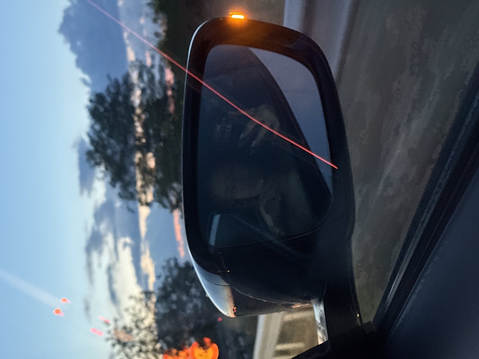 The image shows a side-view mirror of a car. Reflected in the mirror are trees, a sky with clouds and the setting sun with red light, as well as a vague reflection of a person inside the car. The mirror has an integrated orange turn signal. Part of the car window and door frame are also visible. The location is unknown.