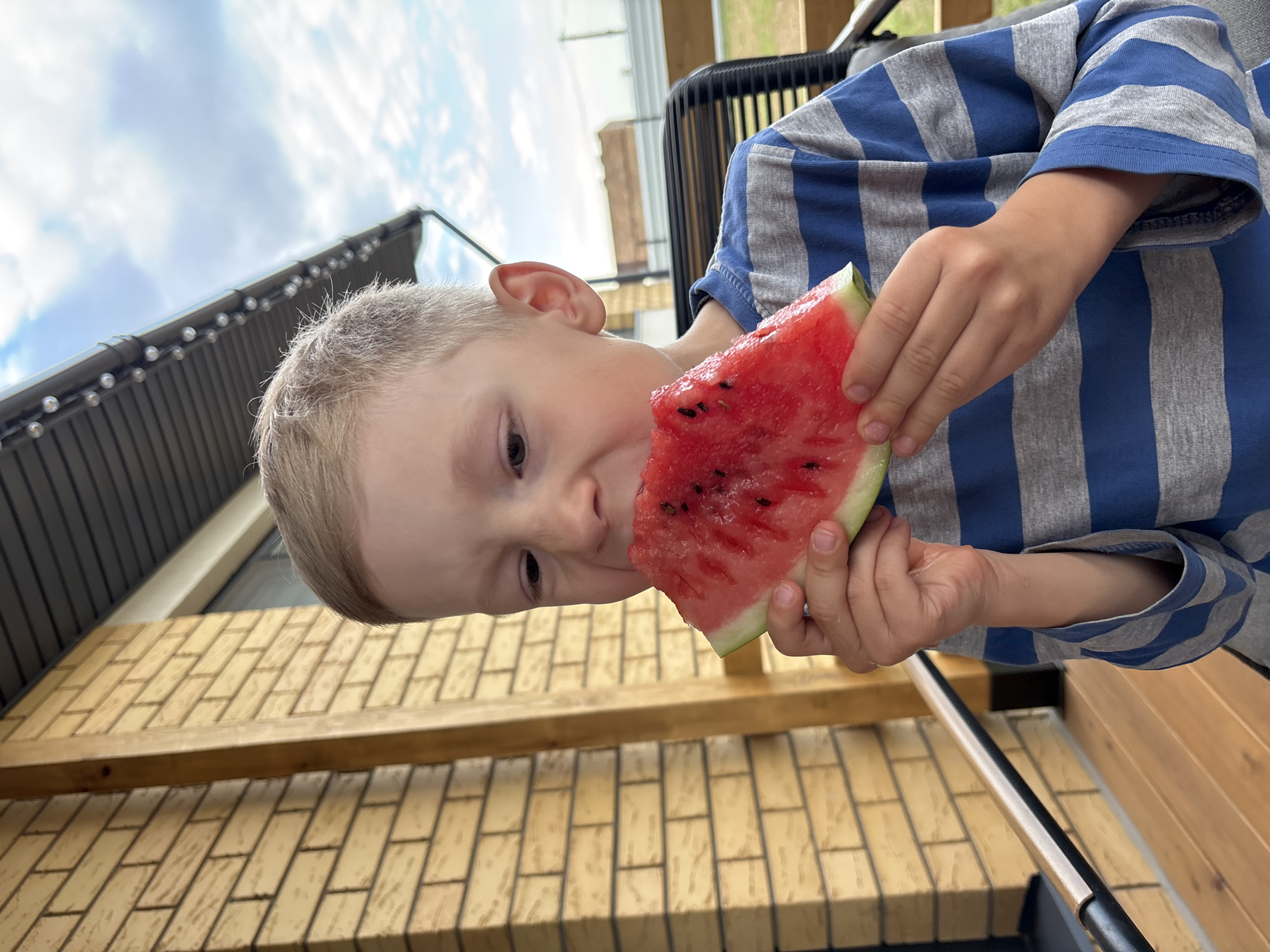 A young boy with blond hair, wearing a blue and gray striped shirt, is sitting on a wooden deck and eating a slice of watermelon. He is holding the watermelon with both hands and taking a bite. The background shows a brick wall and a section of a roof with lights. The boy's eyes are focused on the watermelon.