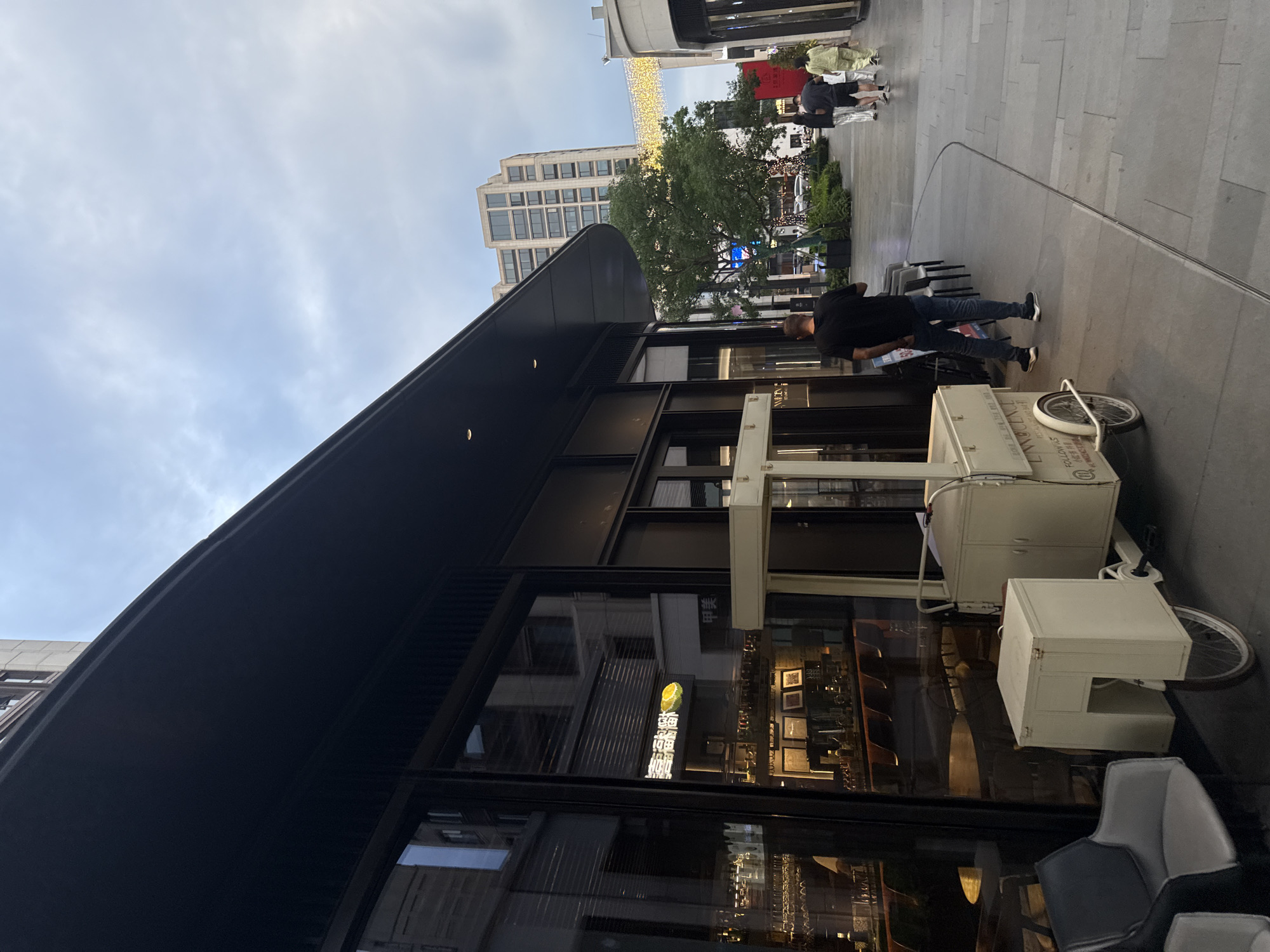 The image shows an outdoor pedestrian walkway in the city of Yiwu, China. The walkway is lined with modern buildings with large glass windows. On the left, a dark-colored awning extends from a building, casting a shadow over the entrance. Inside the building, a bar area is visible through the glass, with bottles and glassware on display. In front of the building, there is a vintage-style white cart with wheels, resembling an ice cream cart or a vendor's stall. A person wearing a black t-shirt and jeans is walking away from the camera down the walkway. Further down the walkway, a group of people, including a man and two women, are walking towards the camera. In the background, a tall, multi-story building with many windows stands out. Decorative lights are strung across the walkway between buildings. Lush green trees are also present in the scene, adding to the urban landscape. The sky is overcast, indicating a cloudy day.