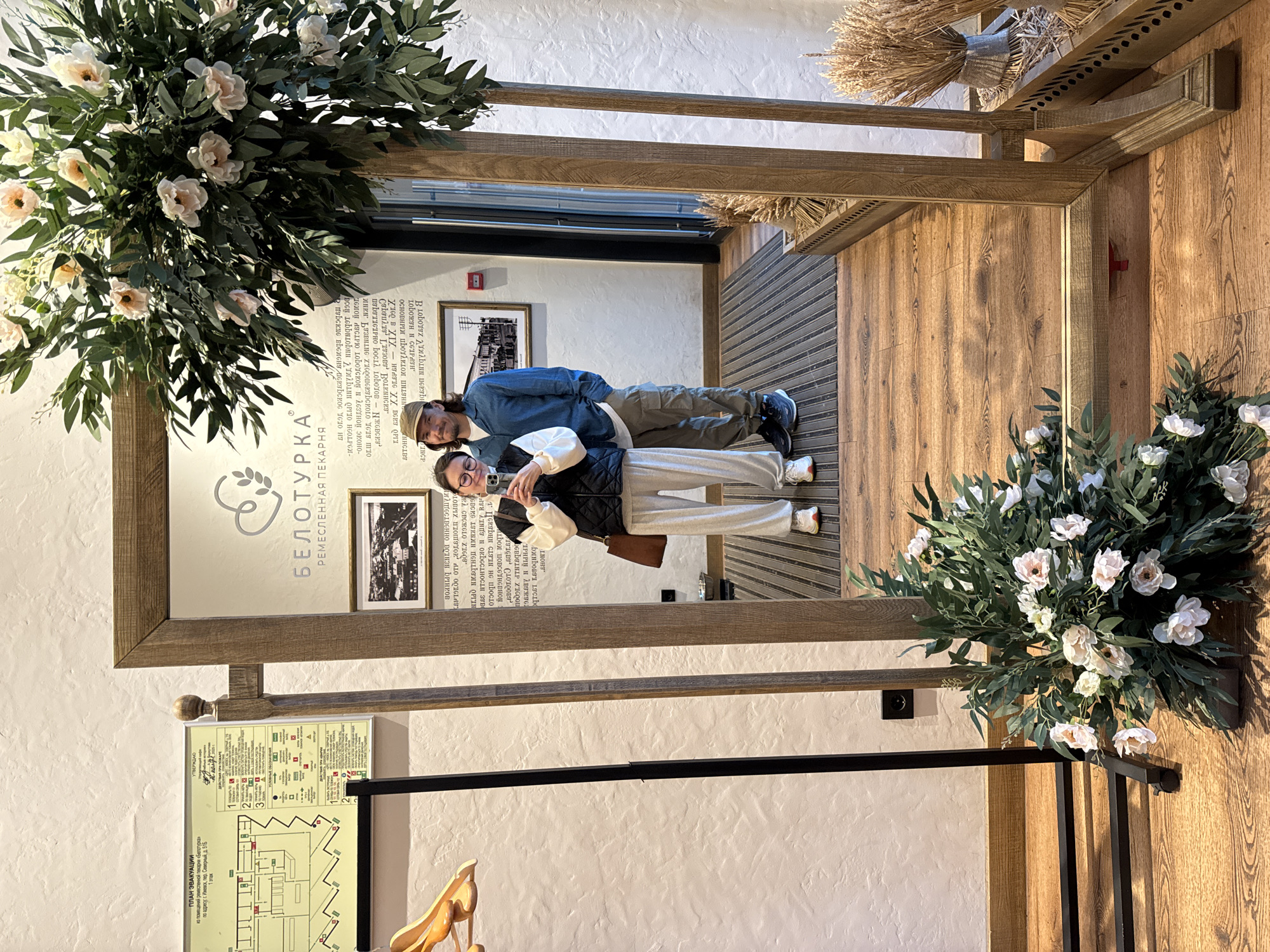 A mirror selfie taken by a female subject captures her and a male subject within the "Beloturka" craft bakery, located at Severny Lane, 51B, Izhevsk, Russia. The female, positioned on the left of the reflection, wears glasses, a black quilted vest over a white long-sleeved top, light grey sweatpants, and white sneakers with red accents, holding a smartphone to take the photograph. A brown shoulder bag hangs from her left shoulder. The male, standing to her right, wears a light brown baseball cap, a blue long-sleeved shirt, olive green cargo pants, and dark sneakers. Both individuals display slight smiles.
The central object is a large, full-length mirror with a substantial, light-brown wooden frame. Artificial floral arrangements, featuring white and pale pink blossoms with green foliage, are attached to the upper right corner of the mirror and placed on the wooden floor in front of its lower left corner.
The reflection reveals elements of the bakery's interior. Above the subjects, the bakery's logo, a heart containing a wheat stalk, is visible with the text "БЕЛОТУРКА" and "РЕМЕСЛЕННАЯ ПЕКАРНЯ." Below this, a framed black and white photograph of an old cityscape hangs on the wall, and a red fire alarm pull station is visible to the left of the male's reflected head.
In the actual room, to the left of the mirror, a white, textured wall features a mounted evacuation plan for the "Beloturka" bakery, confirming its Izhevsk address. A black metal garment rack with a wooden hanger is positioned further left. The floor consists of light-colored wooden planks, and a dark, ribbed entrance mat is partially visible in the reflection behind the male. To the right of the mirror, a wooden decorative container holds dried wheat stalks. An electrical outlet is visible on the wall behind the floor-level floral arrangement.