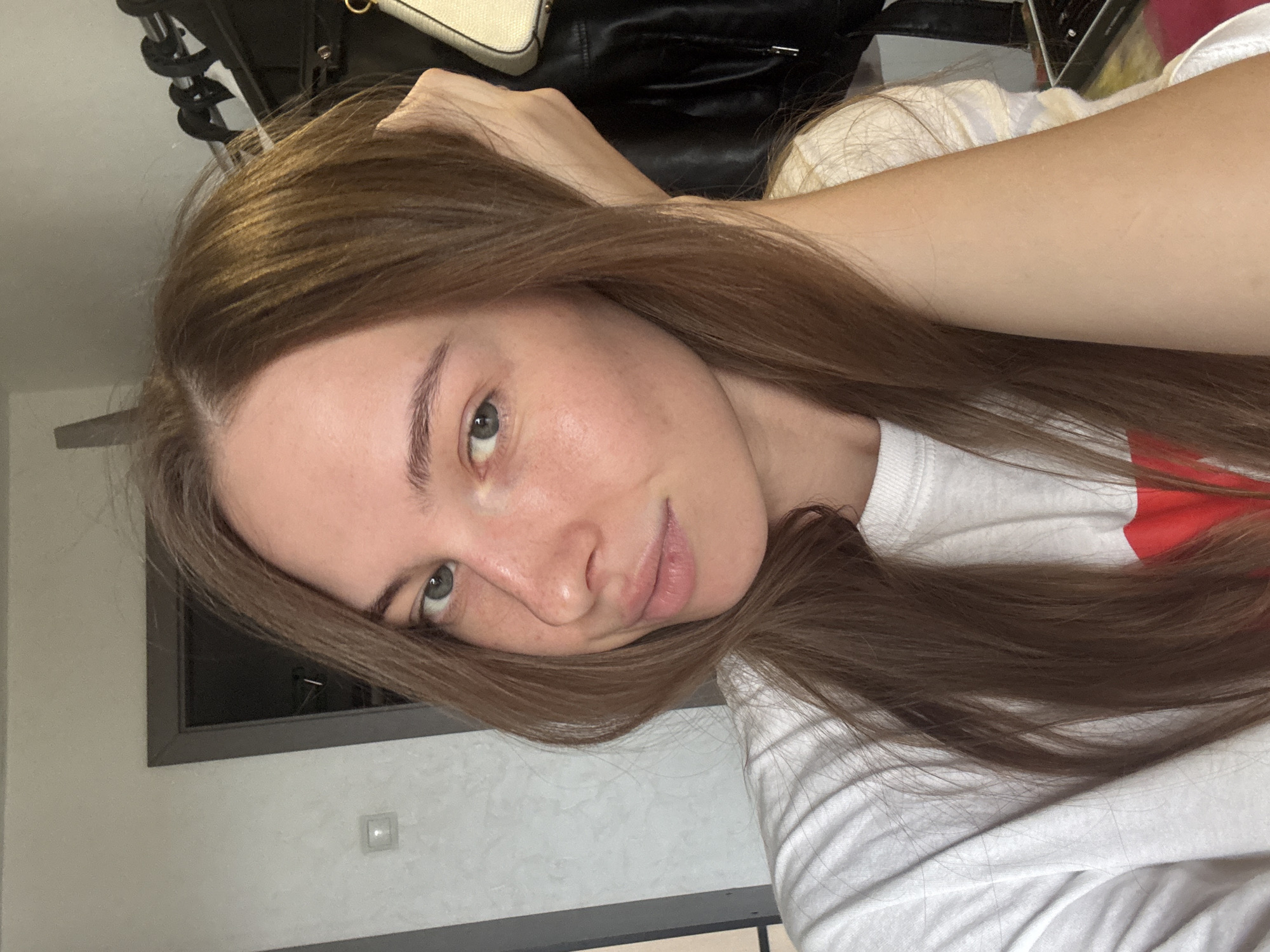 A young woman with long, brown hair and light blue eyes is looking at the camera. She is wearing a white t-shirt with a red design on it. Her right hand is resting on her head, and her hair is falling over her arm. The background is blurry, but appears to be an indoor setting, possibly a room with a door and a closet. There is no indication of the city of Perm, Russia.