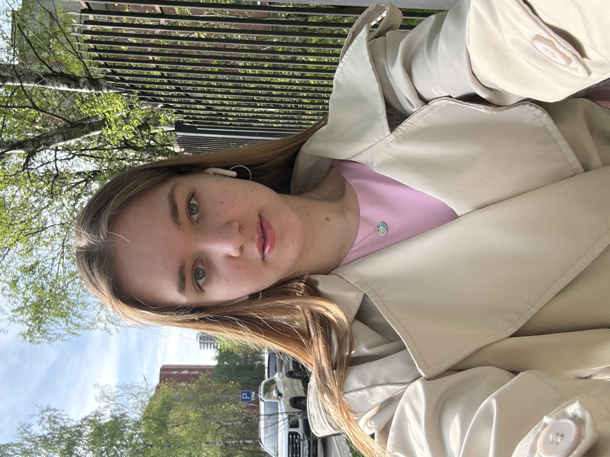 A young woman with light brown hair, green eyes, and fair skin takes a selfie. She wears a pink top and a beige trench coat. She has wireless earbuds in her ears and a necklace. Behind her is a black metal fence, trees with green leaves, buildings, and parked cars. A parking sign is visible. Based on the background, the image appears to be taken in an urban area, possibly the city of Perm, Russia.