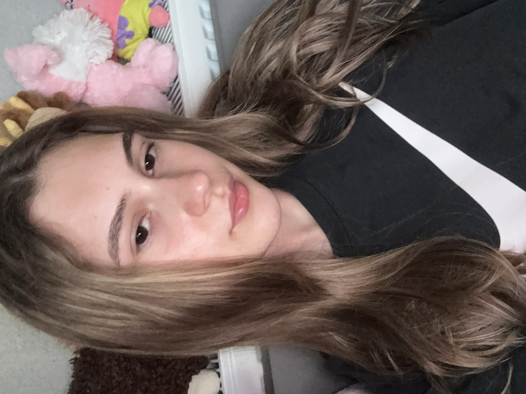 The image shows a young woman lying down, possibly on a bed, with plush toys visible near her head. She has light brown hair with blonde highlights and is wearing a black t-shirt with a white Nike logo. The background is blurred, making it difficult to discern any specific details about the environment. There is no information available in the image to indicate the location is Panagyurishte, Bulgaria.