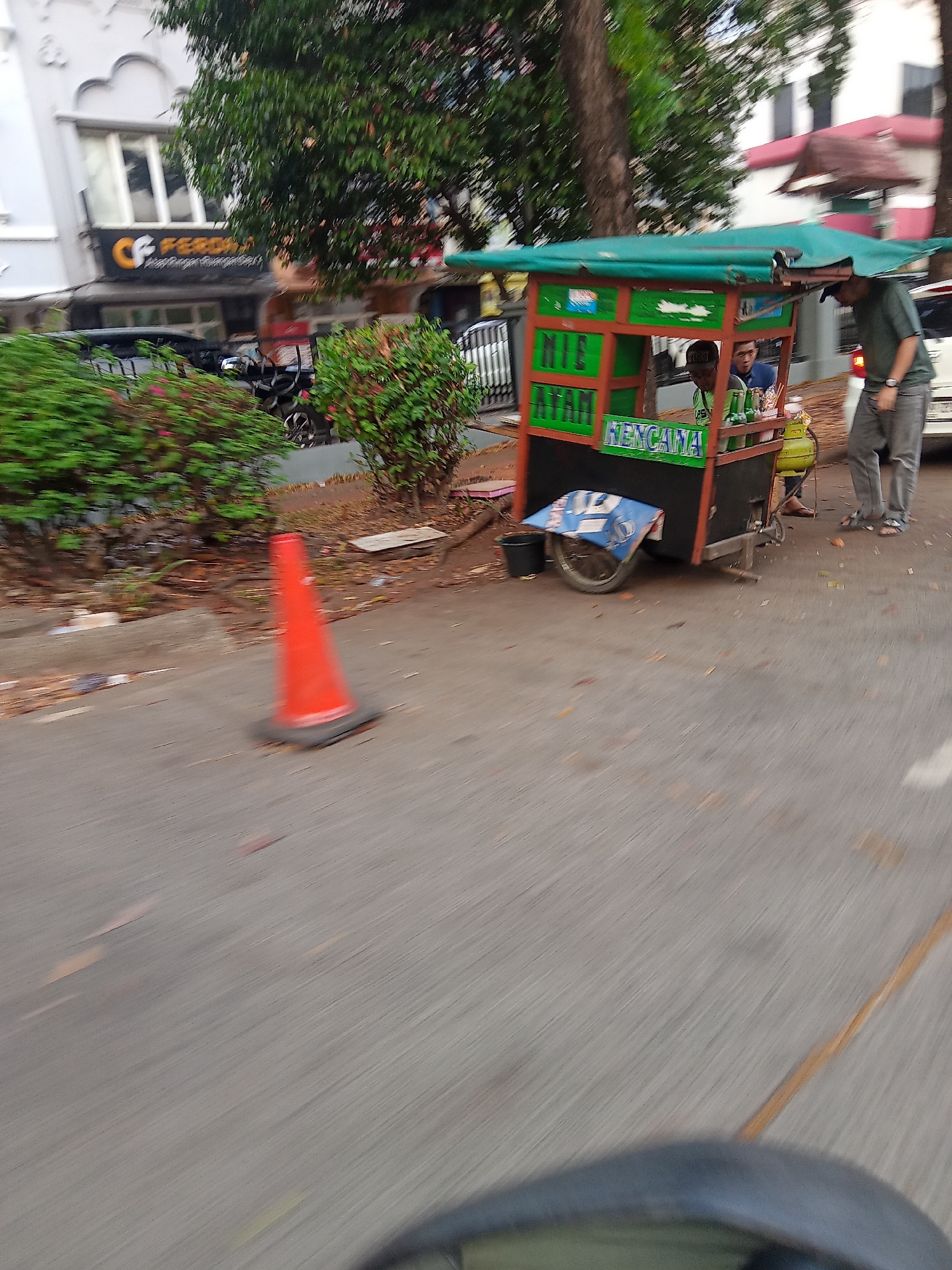 An outdoor, slightly blurred scene depicts a food cart with a green awning in front of a building with a prominent signage that says "CF Feronia". The cart has "MIE AYAM RENCANA" written on its side. Two men are near the cart; one appears to be working, and the other is standing behind him. A red traffic cone is on the road in the foreground. The environment suggests a street in the city of South Tangerang, Indonesia, possibly during daylight hours.