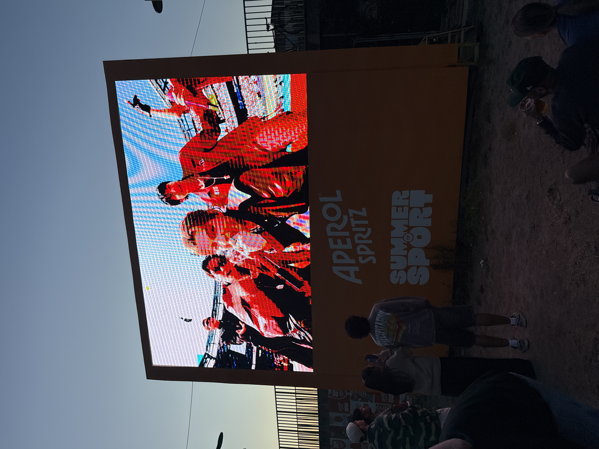 A large LED screen displays a video of a group of young people, possibly musicians, in red shirts, with one person in the foreground wearing a necklace and a black shirt. The screen is mounted on an orange structure with the words "APEROL SPRITZ SUMMER OF SPORT" visible below the video. In the foreground, several people are watching the screen, some standing and some sitting. The ground is covered in dirt and gravel. A dark fence and a bicycle are visible in the background on a raised platform to the right of the screen. The sky above is a pale blue. The overall setting appears to be an outdoor event.