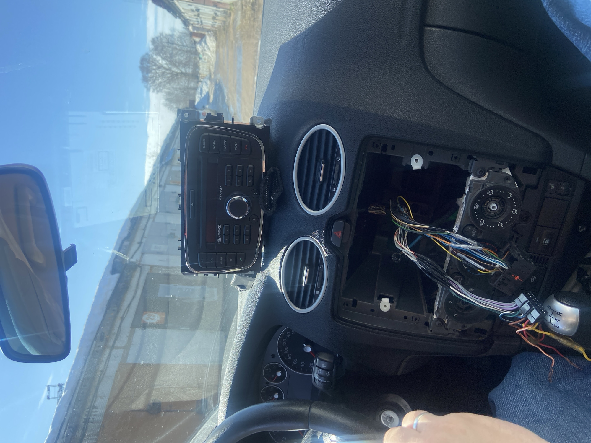 The image, taken from within a vehicle, shows the front passenger compartment of a car with a partially disassembled dashboard. A Ford 6000 CD radio unit, labeled "CD," "AUX," and "FM/AM," is detached and resting on the dashboard, connected by exposed wiring. Below its original position, the central console cavity is open, revealing a tangle of multi-colored electrical wires and connectors. To the left, a portion of the steering wheel and the instrument cluster are visible, with the speedometer needle indicating a speed below 20 km/h. A driver's hand, wearing a ring, is visible on the left side, near the steering column. Oval-shaped air vents with chrome rings are prominent on the dashboard, alongside a red hazard light button. A manual gear shifter is visible below the exposed wiring.
Outside the car, through the windshield, a bright, clear blue sky is visible. The vehicle is situated on an unpaved road flanked by rows of single-story, light-colored brick or concrete garage-like buildings or workshops. These buildings have white roofs, partially covered with snow, and piles of snow line the sides of the road. One building on the left features a red circular sign with the number "29" inscribed. Further down the road on the right, numbers "21" and "20" are discernible on another building. Bare trees are visible in the distance beyond the structures. This environment is consistent with a garage cooperative or industrial area typically found in the outskirts of a city such as Chaikovsky, Russia.