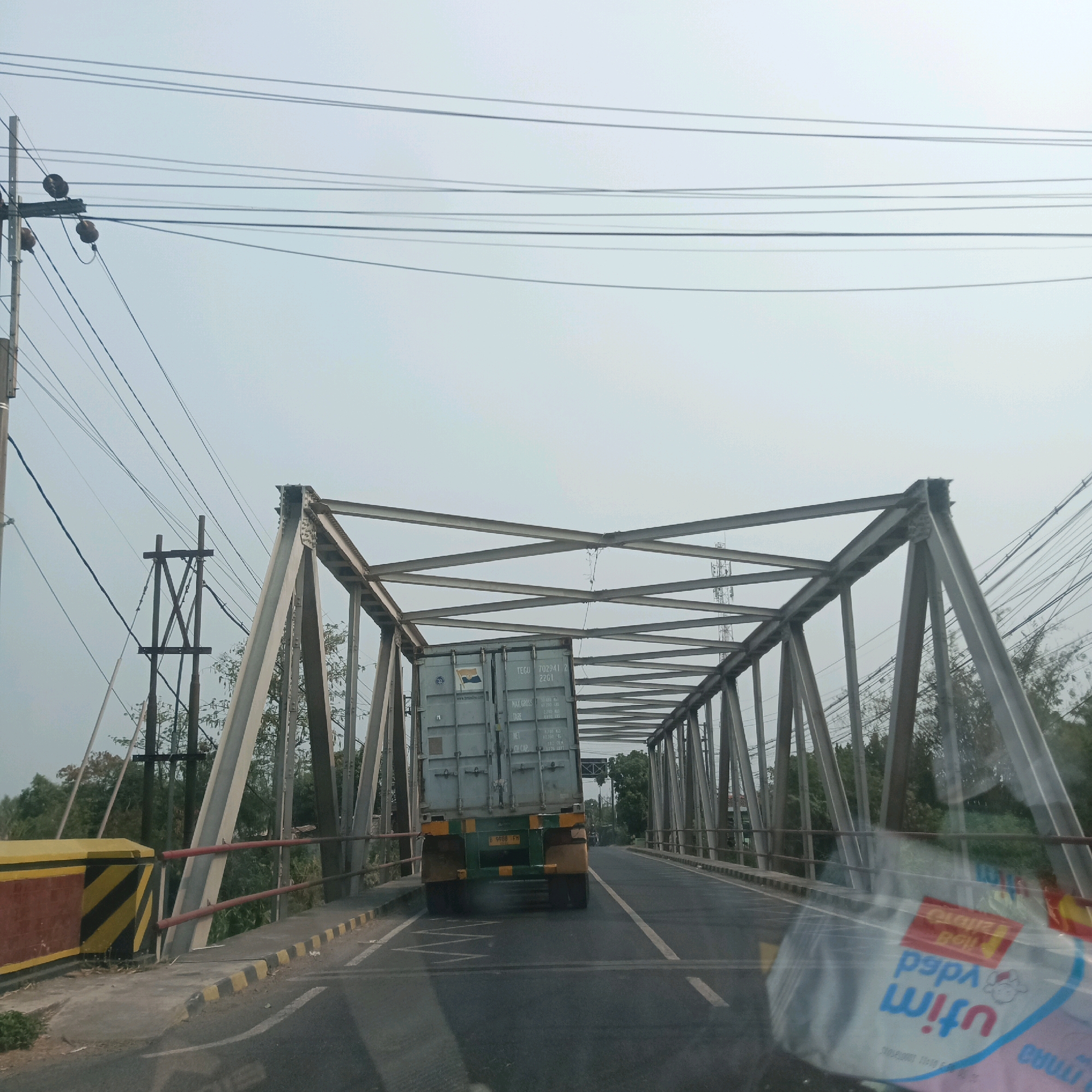 The image captures a view from a vehicle traveling across a metal truss bridge in Jetis, Indonesia. The bridge has a gray metal structure with diagonal supports. A large, light-blue shipping container is being transported by a truck on the bridge. The truck's license plate reads "B 9098 FH". Power lines run overhead, supported by utility poles. The bridge deck is asphalt with yellow and black striped markings on the side. Trees and foliage are visible on either side of the bridge. The sky is overcast and pale. In the foreground, a plastic water bottle is partially visible on the dashboard of the vehicle, with a distorted reflection of the scene on its surface.
