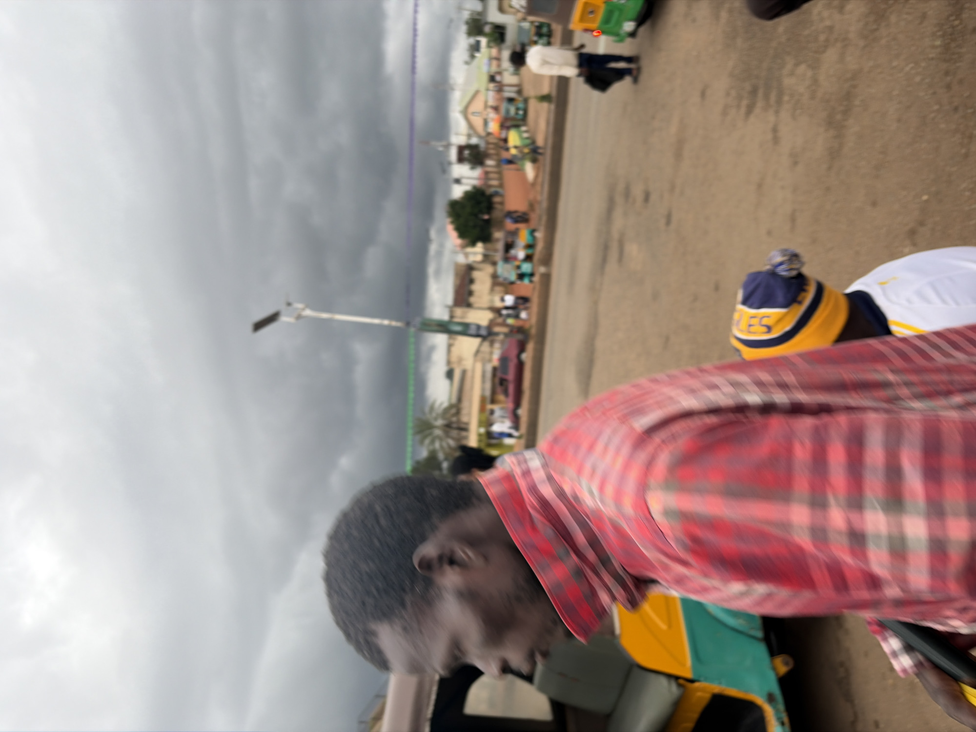 In Gura Top, Nigeria, under a cloudy sky, a man in a red and black plaid shirt stands near a yellow and green auto-rickshaw. To his right, another person wears a blue and yellow striped hat. In the background, several other auto-rickshaws and a few individuals are visible on a paved road. Buildings and palm trees line the street, with a lamppost in the distance.
