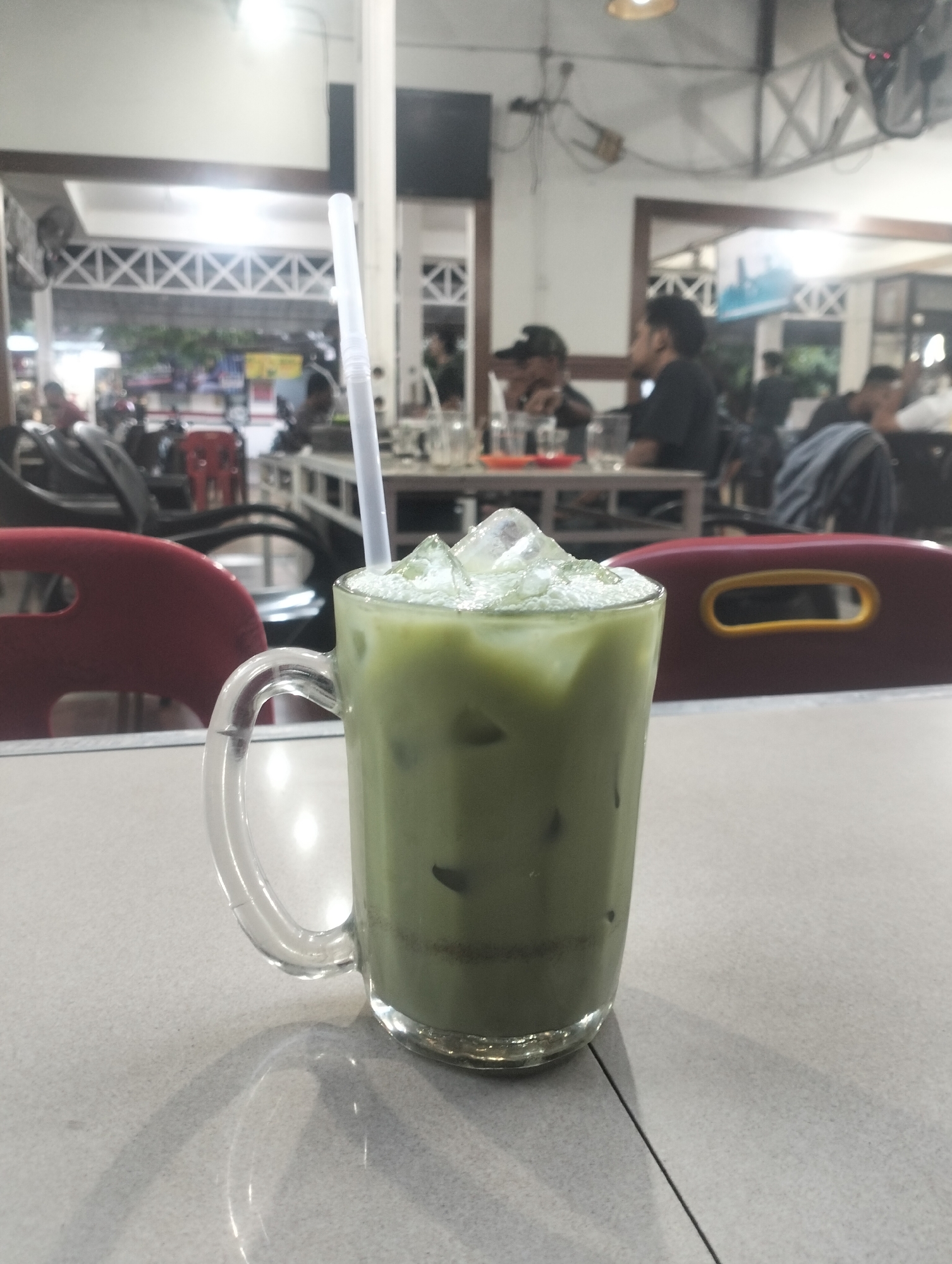 A green drink with ice in a glass mug sits on a light gray table. A white straw protrudes from the drink. Red chairs are visible behind the glass. In the background, people are seated at tables within a restaurant-like setting. A ceiling fan is visible on the right, attached to the ceiling. The location appears to be a cafe or restaurant in Lhokseumawe, Indonesia.