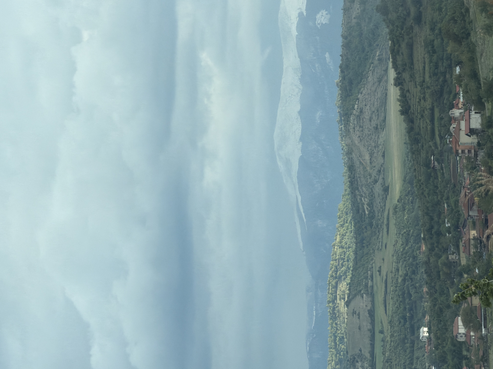 The image presents a serene, expansive mountainous landscape under an overcast sky. The scene is horizontally oriented, with the sky occupying the left portion of the frame and the terrestrial elements extending from the middle to the right.
The sky is dominated by thick, diffuse clouds in varying shades of light blue and grey, suggesting a cloudy day with soft, indirect lighting.
To the right, a dramatic panorama unfolds, starting with distant, towering mountains whose peaks appear rugged and lighter in color, possibly due to bare rock or a thin dusting of snow. These high peaks are partially obscured by atmospheric haze, blending into the cloudy sky.
Closer to the viewer, a prominent, lush green forested mountainside or hill runs across the middle ground, covered in dense, dark green trees. Below this forested area, patches of lighter green fields or pastures are visible, indicating open land or agricultural use.
In the right foreground, nestled into the landscape, is a small, picturesque village. Numerous houses with reddish-brown tiled roofs and predominantly light-colored, likely white or cream, walls are scattered across a gentle slope. The buildings are interspersed with trees and greenery, integrating harmoniously with the natural surroundings.
The image captures a tranquil, rural setting, devoid of any visible human or animal activity. The time of day is daytime, characterized by diffused light due to the heavy cloud cover. There is no discernible text within the image. The landscape evokes a sense of peace and natural beauty, consistent with a remote area such as Belo Pole, Bulgaria.