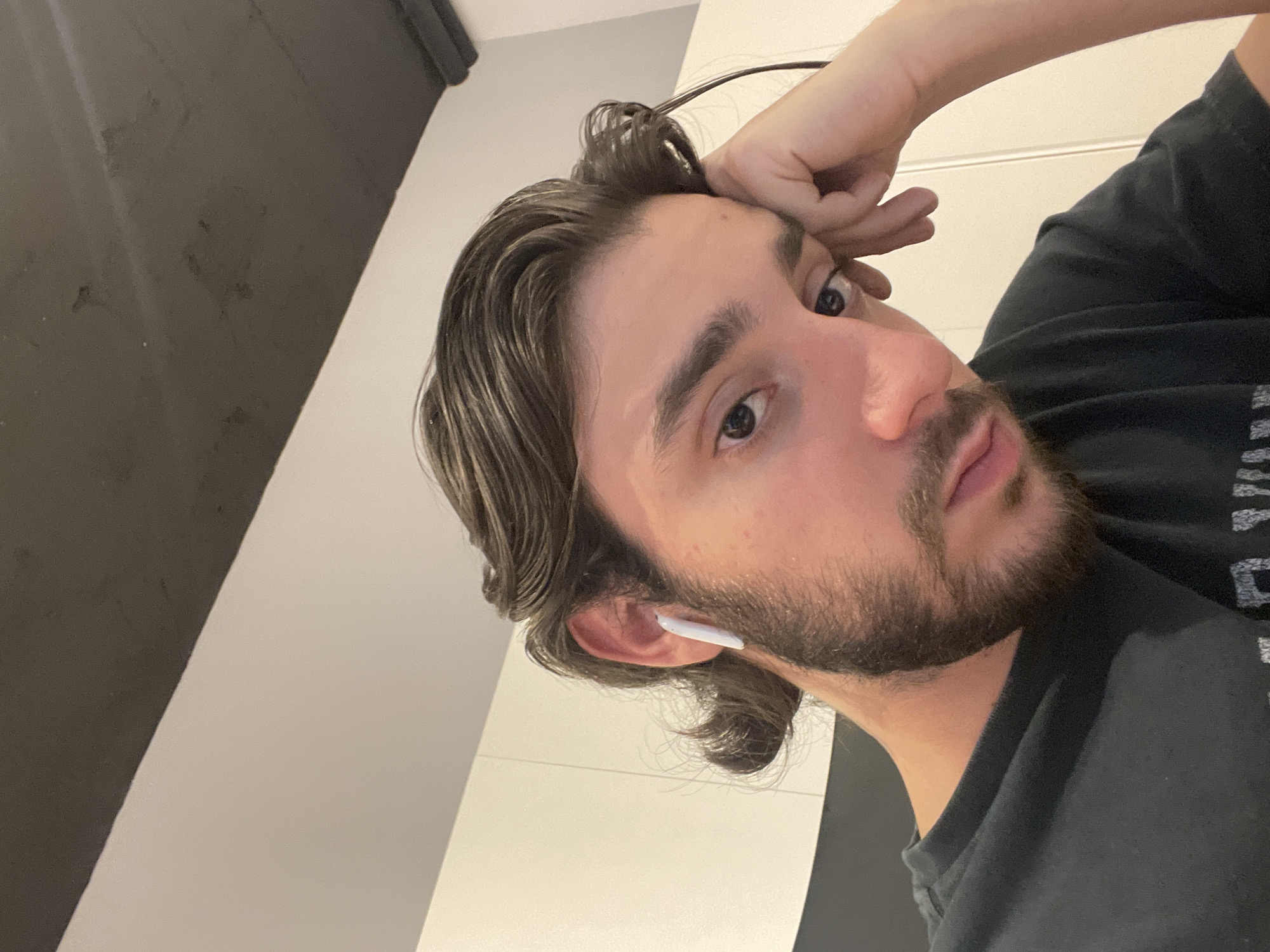 A close-up, slightly angled, selfie of a person with brown hair and a beard, wearing a black t-shirt and a white wireless earbud in their left ear. The background consists of a light-colored wall and a dark-colored wall or object, possibly a ceiling. The location is unknown and there is no clear indication that the image was taken in Mexico City.
