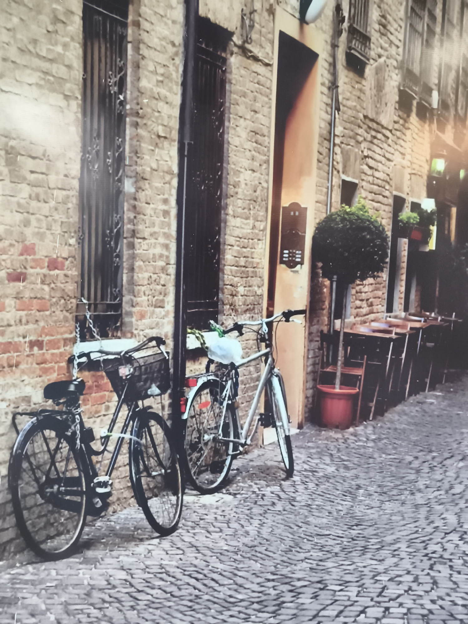 The image presents a European street scene, not one from Baan Bo Phut, Thailand. Two bicycles are parked against a brick building. The building features barred windows and a doorway, along with outdoor seating. The street is paved with cobblestones. No people are clearly visible, but there are tables and chairs suggesting a restaurant.