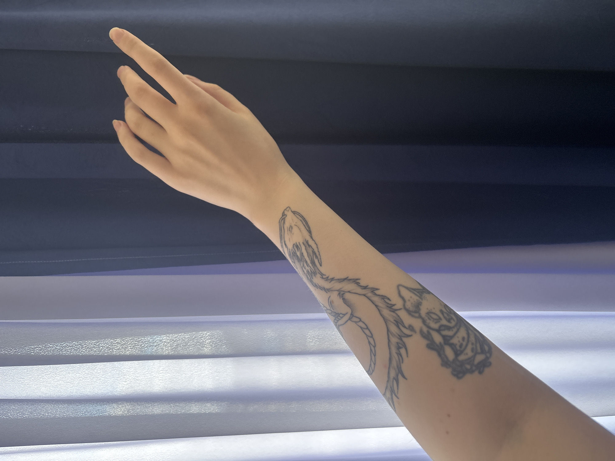 An arm with a detailed black ink tattoo is shown extending upwards against a backdrop of horizontal blinds. The tattoo seems to depict fantasy creatures and designs. The skin of the arm is light-toned. The blinds alternate between dark blue and a light grey-white, creating contrasting bands of color behind the arm. There is no evidence of this image being connected to the city of Верхнеказымский, Russia.
