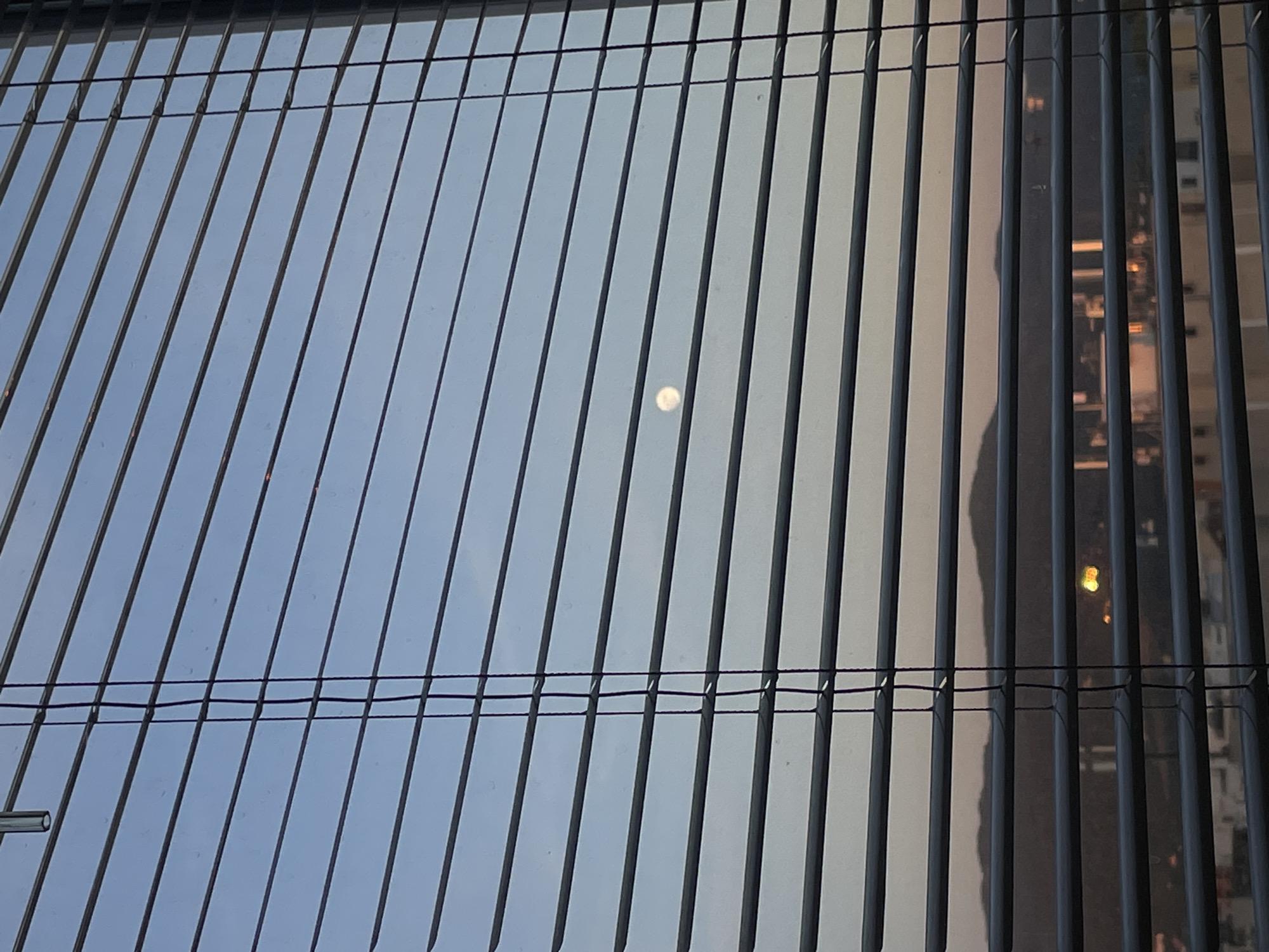The image shows a view through closed vertical blinds. Outside, a light source, possibly the moon or a bright light, is visible between the slats. The background on the right side of the image reveals blurred cityscape details, potentially buildings and lights, suggesting the location is urban. The color and brightness of the sky, glimpsed between the blinds, suggest twilight or early evening. The scene does not depict any people. It is difficult to determine from the image if this is Sofia, Bulgaria, without additional context or clearer visual cues.