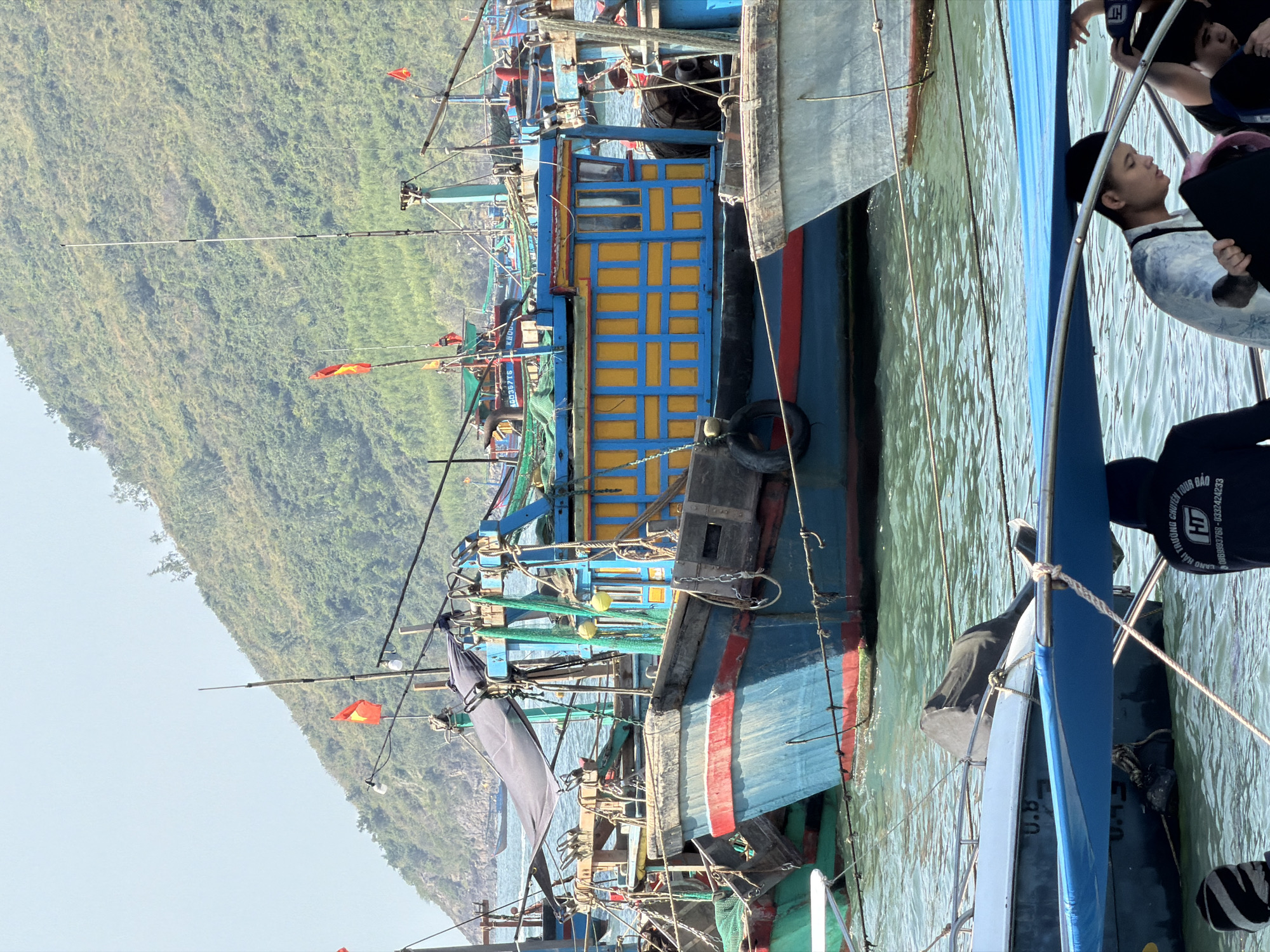 The image shows several fishing boats docked in Nam Nha Trang Ward, Vietnam. The boats are various colors and designs, with some displaying the Vietnamese flag. A lush green hill is visible in the background. People are onboard at least one of the boats.