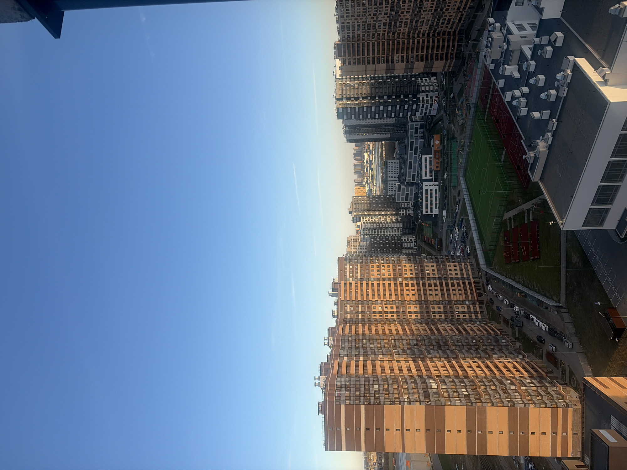 The image presents a high-angle view of a dense urban residential area, likely taken from an upper floor of a tall building. The scene is dominated by numerous high-rise apartment buildings under a clear, bright blue sky with a few faint contrails.
The architecture features a mix of modern designs, with some buildings exhibiting dark grey or black facades and others a warm, light brown or orange hue, suggesting varied construction materials or paint. Many windows are visible across the building exteriors. The buildings are tightly packed, indicating a high-density urban development.
At ground level, several amenities are visible. A large green sports field, likely a soccer pitch, with white markings is clearly depicted, adjacent to a smaller reddish-brown court. Roads wind through the complex, with numerous parked cars lining the streets. Some green spaces and trees are interspersed between the buildings and facilities. The roofs of closer buildings show an array of ventilation units and other rooftop infrastructure.
The lighting suggests late afternoon or early morning, with the sun casting long shadows and illuminating some building facades with a warm, golden glow, contrasting with the shadowed sides. The weather is clear and sunny. There are no clearly discernible people or specific activities taking place, nor is there any visible text on buildings or signs. The overall impression is one of a modern, well-planned residential district in a large city like Saint Petersburg, Russia.