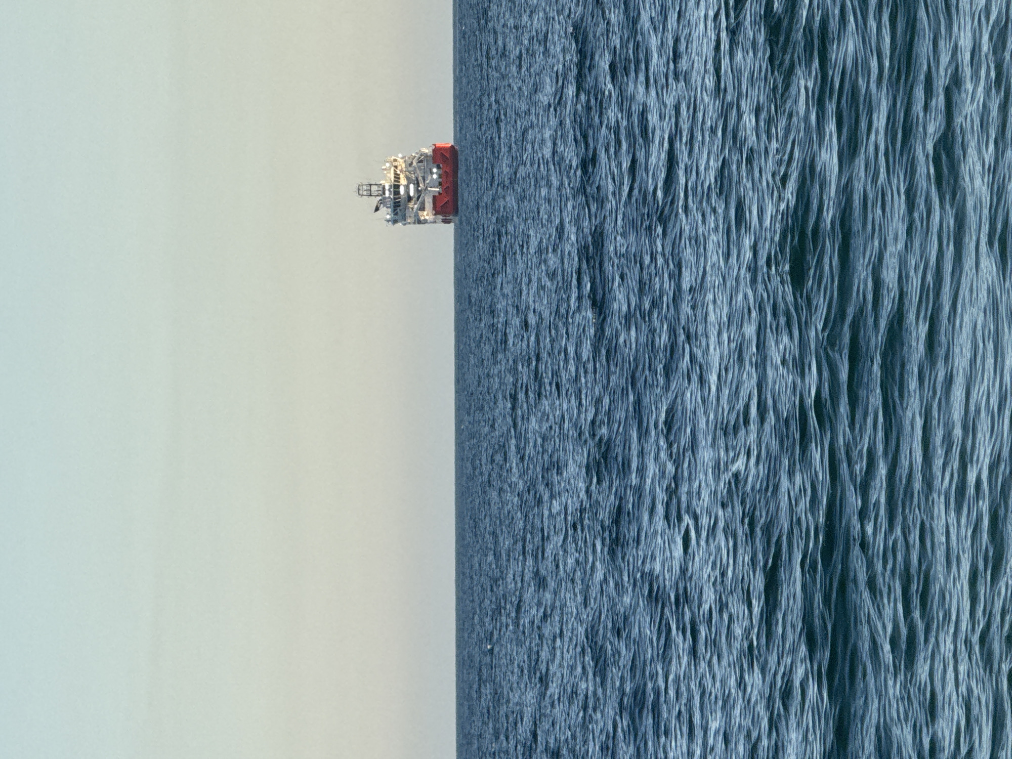 A large vessel with a red hull and white superstructure is seen at sea, positioned on the horizon. The vessel is rectangular in shape, with multiple decks and structures on its upper portion. The water in the foreground is a deep blue, with visible waves and ripples. The sky above is a pale, gradient color, transitioning from a light blue at the top to a yellowish hue near the horizon. There are no people visible in the image. The scene appears to be an open body of water, potentially off the coast of Niterói, Brazil, given the typical landscape of Guanabara Bay.
