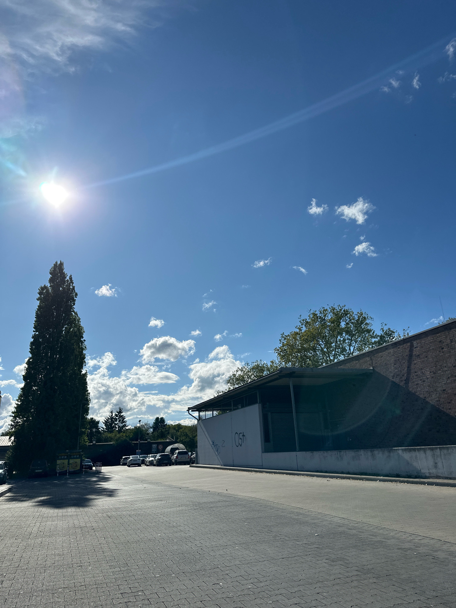 The image depicts an outdoor scene in Krefeld, Germany, on a clear, sunny day with scattered clouds. The foreground is a paved area made of interlocking grey bricks, suggesting a parking lot or a pedestrian walkway. To the left, a tall, slender evergreen tree stands casting a shadow. Behind the tree, several cars are parked in a lot. Further back, there are more trees and some low-lying structures, possibly sheds or industrial buildings.
Dominating the middle ground is a building with a white facade, partially covered by a dark, sloped roof. This building has some graffiti on its white wall, including the characters "Z. OST". To the right of this building, a dark brick wall rises, and further to the right, more greenery and sky are visible.
The sky is a vibrant blue with several white, fluffy clouds. The sun is shining brightly from the upper left quadrant of the image, creating lens flares and streaks of light across the sky and parts of the scene. No people are visible in the image. The environment suggests a commercial or industrial area within the city of Krefeld.