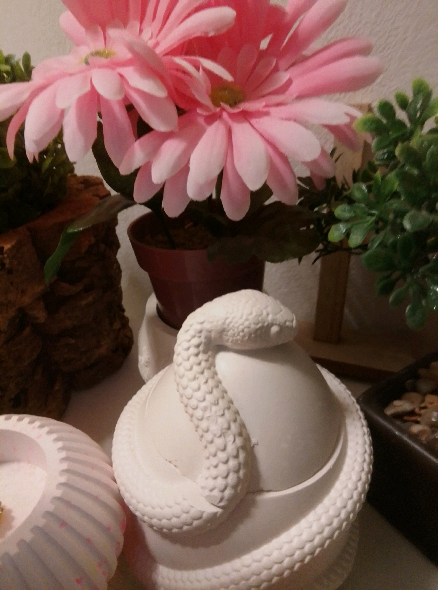 The image depicts a still life arrangement with a ceramic snake coiled around a white, lidded pot in the foreground. Behind the pot, there is a cluster of artificial pink flowers with visible pistils and green foliage. To the left of the pot, a textured, bark-like object is partially visible, and to the left of that, a fluted, white cylindrical object with colorful flecks is present. To the right of the pot, a dark rectangular container filled with small stones is visible. The background is a plain, textured white wall. The scene appears to be indoors, likely on a surface such as a table or shelf, with soft lighting. There are no people or discernible actions taking place. The location in the city of Wedel, Germany is not indicated by any visual cues in the image.