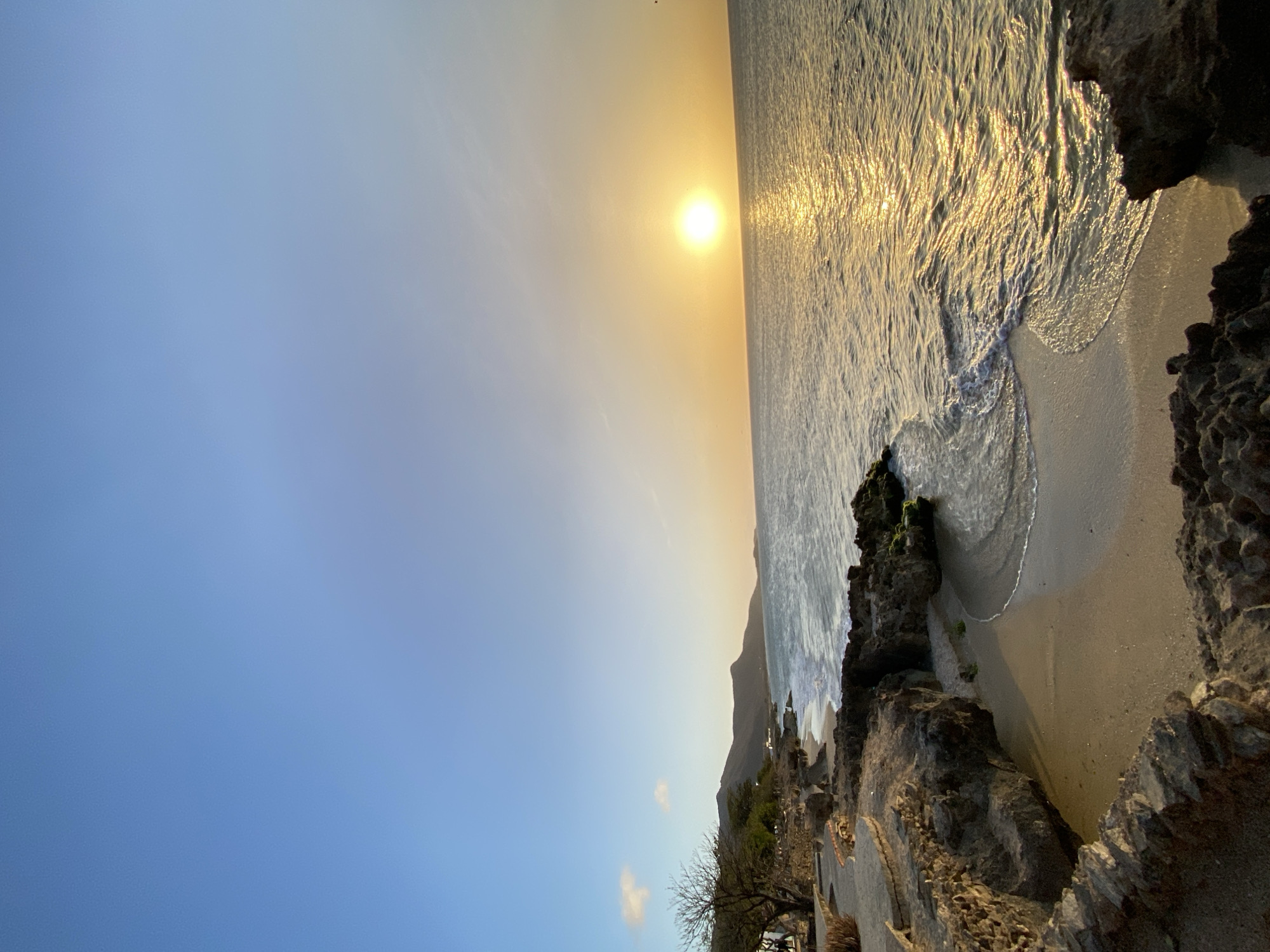 The image shows a coastal scene in Juan Griego, Venezuela at what appears to be sunset. The sun is positioned on the horizon over the ocean, casting golden reflections on the water's surface. The sky transitions from a bright blue in the lower left to a warmer yellow-orange near the horizon. The shoreline is rocky, with dark, jagged rocks framing a small sandy beach. Waves are lapping at the shore. Some sparse vegetation is visible along the rocky coastline, further inland. There are a few buildings visible in the distance.