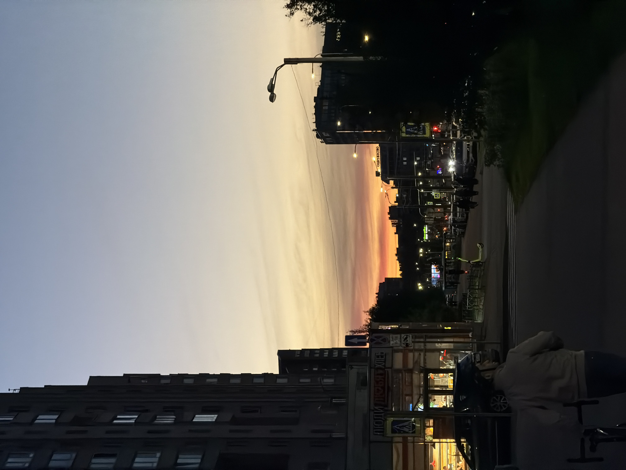 The image depicts a street scene in Saint Petersburg, Russia, during a sunset. A tall, dark apartment building dominates the left side of the frame. Across the street, a row of shops is visible, with illuminated windows and signage. Further down the street, more buildings and streetlights are illuminated, creating a warm glow against the dusky sky. The sky itself transitions from a pale blue at the top to vibrant oranges and reds near the horizon, indicating the setting sun. Several cars are parked and driving on the street. A person wearing a light-colored jacket and a helmet is walking away from the camera on the sidewalk. The overall atmosphere is urban and twilight.