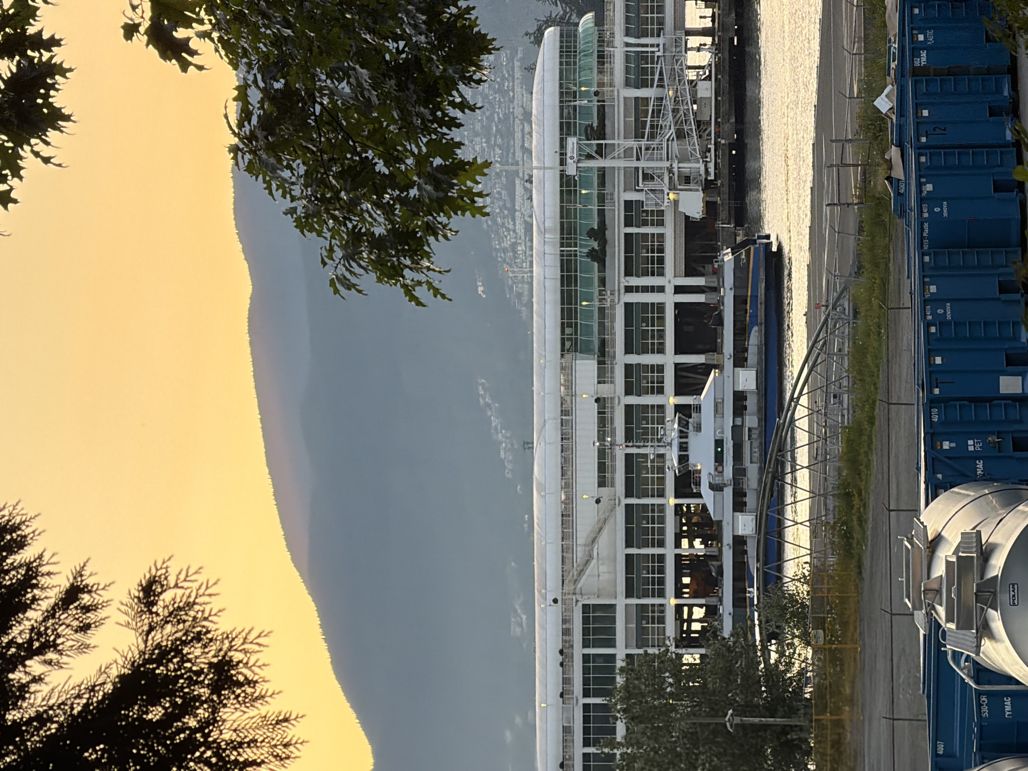 The image depicts a view of the waterfront in Vancouver, Canada, with a large ferry terminal building in the midground. The sky is a warm, hazy yellow, suggesting either sunrise or sunset. In the foreground, several large blue recycling bins are visible, with a silver tank-like container on top of one of them. Beyond the recycling bins, a chain-link fence separates the area from a paved surface and the water.
The ferry terminal building is a multi-story structure with a white upper section featuring many windows and a curved roof. A ferry is docked alongside the terminal, with its ramp extended. The water is calm, reflecting the warm light of the sky. In the background, hazy mountains rise, with some structures visible on their slopes. Trees frame the top and right sides of the image, their leaves silhouetted against the bright sky.
The overall scene is one of industrial and transportation activity at the edge of a natural landscape, bathed in the soft light of dawn or dusk. There are no visible people in the frame.