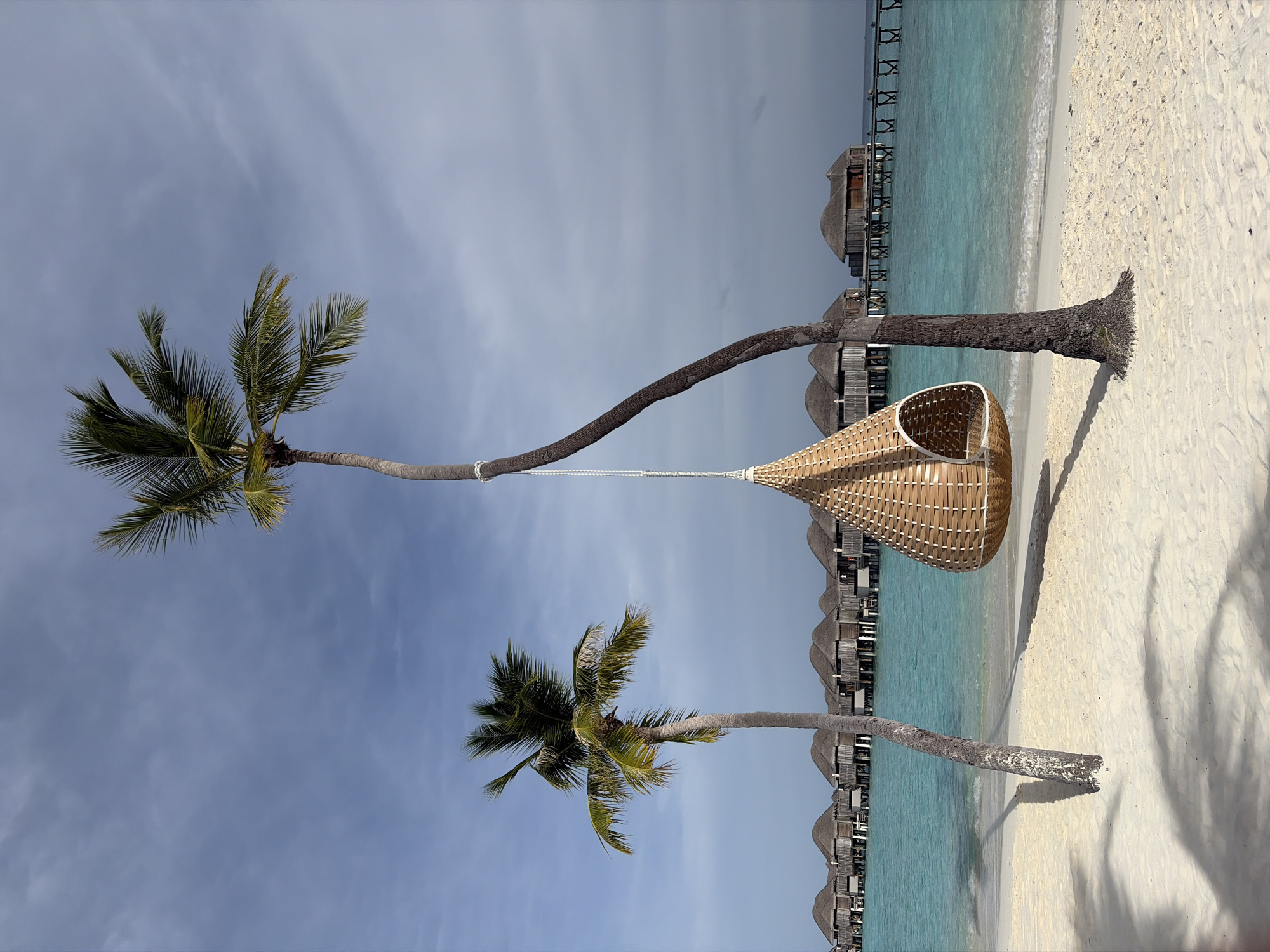 The image depicts a tropical beach scene in Halaveli, Maldives. In the foreground, white sand stretches towards a turquoise ocean. Two palm trees are prominent; one leans precariously to the right, supporting a woven hanging chair suspended by a rope. The other palm tree stands to the left, with its trunk casting a shadow on the sand. In the background, a row of overwater bungalows with thatched roofs lines the shore, connected by wooden walkways. The sky is partly cloudy, with a mix of blue sky and soft white clouds. The scene conveys a sense of serene, idyllic vacation resort atmosphere. There are no people visible in the frame.