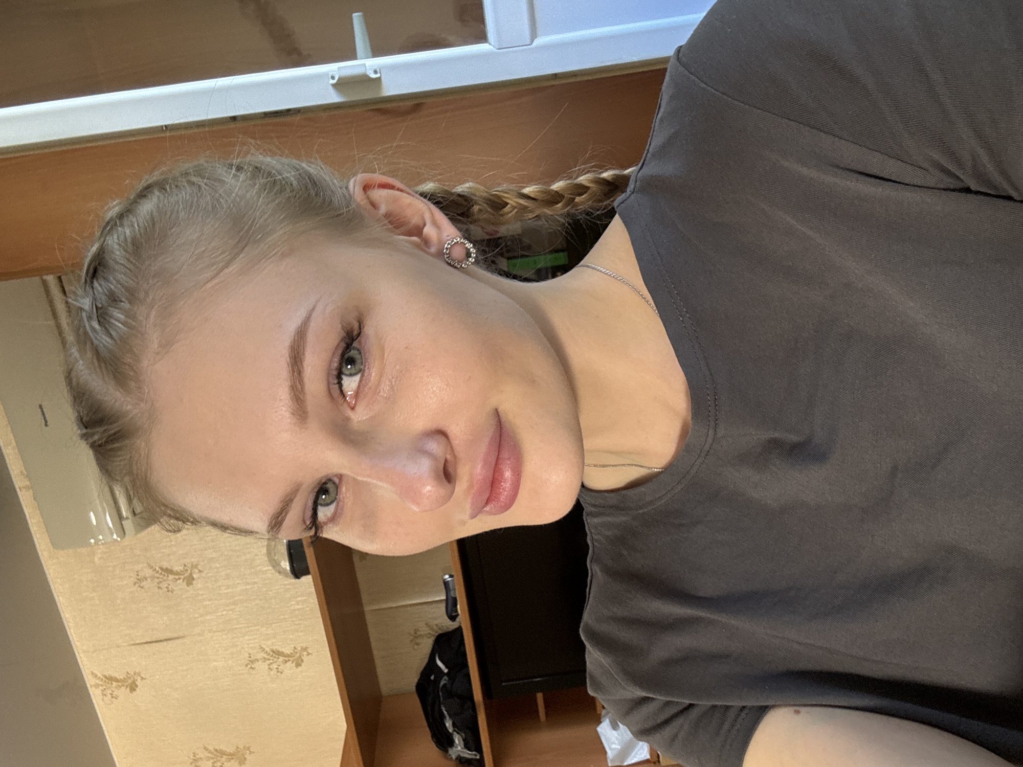 A young woman with blonde hair braided into pigtails and light makeup is taking a selfie indoors. She is wearing a dark gray t-shirt and a silver necklace. She has on circular earrings with small stones. The background shows a wall with floral wallpaper, shelves with clothing and an air conditioning unit. The overall scene suggests a domestic environment, possibly an apartment. The presence of a Panasonic air conditioner unit and the style of the wallpaper could be indicative of a location in Russia, such as Yekaterinburg.