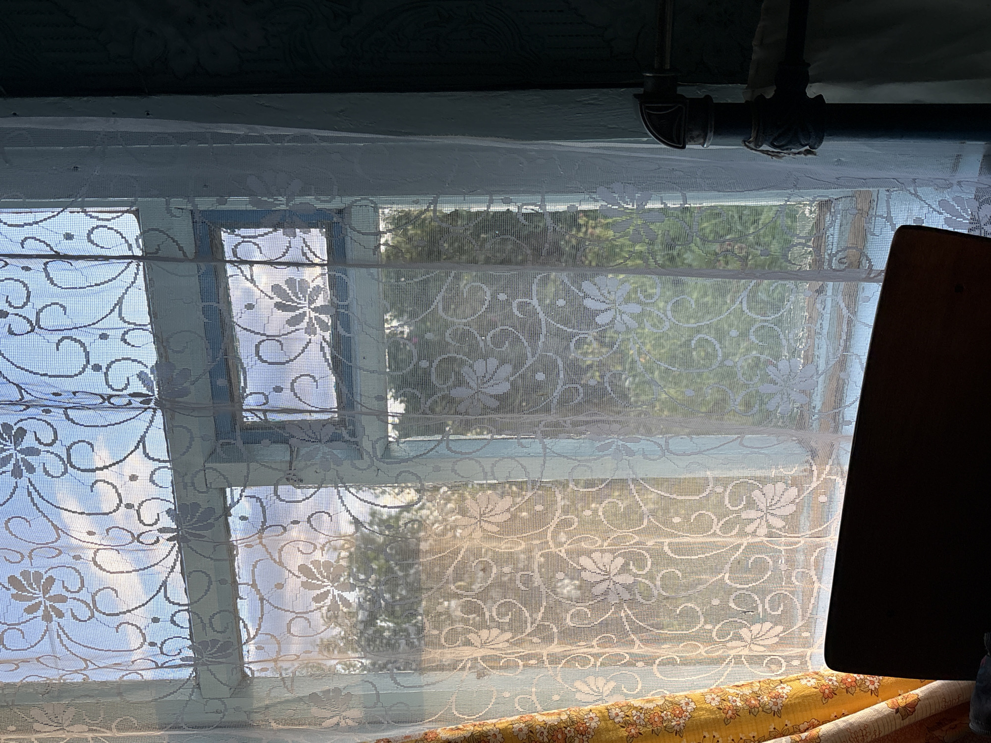 An interior shot taken through a window in Відзаўскі сельскі Савет, Belarus, reveals a view of trees and the sky. The window is draped with sheer white lace curtains featuring a floral pattern and a yellow curtain with a floral print is partially visible on the left. Outside, green trees are seen against a blue sky with white clouds, illuminated by sunlight. Inside, a dark wooden object, possibly a chair back, occupies the lower left portion of the frame. A metal pipe runs vertically along the right side of the image, attached to what appears to be a radiator. The wall to the right is dark and textured.