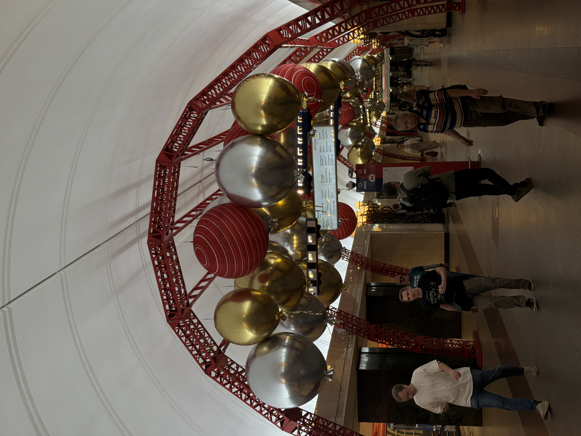 The image depicts the ZIL Moscow metro station. Red steel supports crisscross across the vaulted white ceiling, and oversized Christmas ornaments in gold, silver, and red hang from them. Several people walk on the platform.