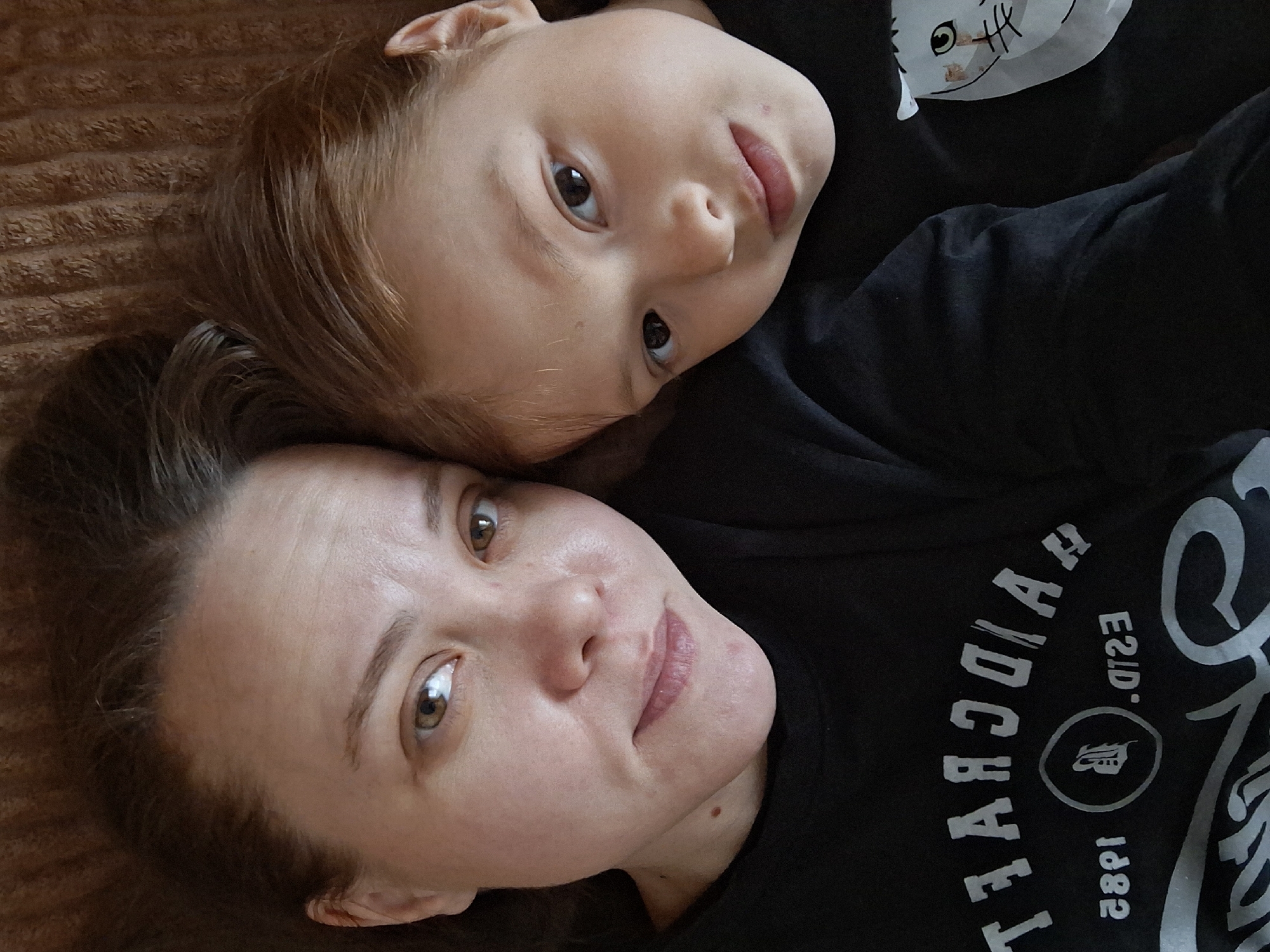 A low-angle, close-up shot shows a woman and a child lying on a brown, textured surface, possibly a bed or couch. The woman is positioned closer to the bottom of the frame and has light skin, brown hair, and brown eyes. The child, also with light skin and brown hair, rests their head near the woman's head. Both are wearing dark-colored clothing. Part of a design is visible on the woman's shirt, featuring the word "HYDE" in a curved arrangement, along with "ESTD." and the year "1882." The environment is otherwise indistinct. The location in the city of Chaikovsky, Russia, cannot be determined from the visual information provided.