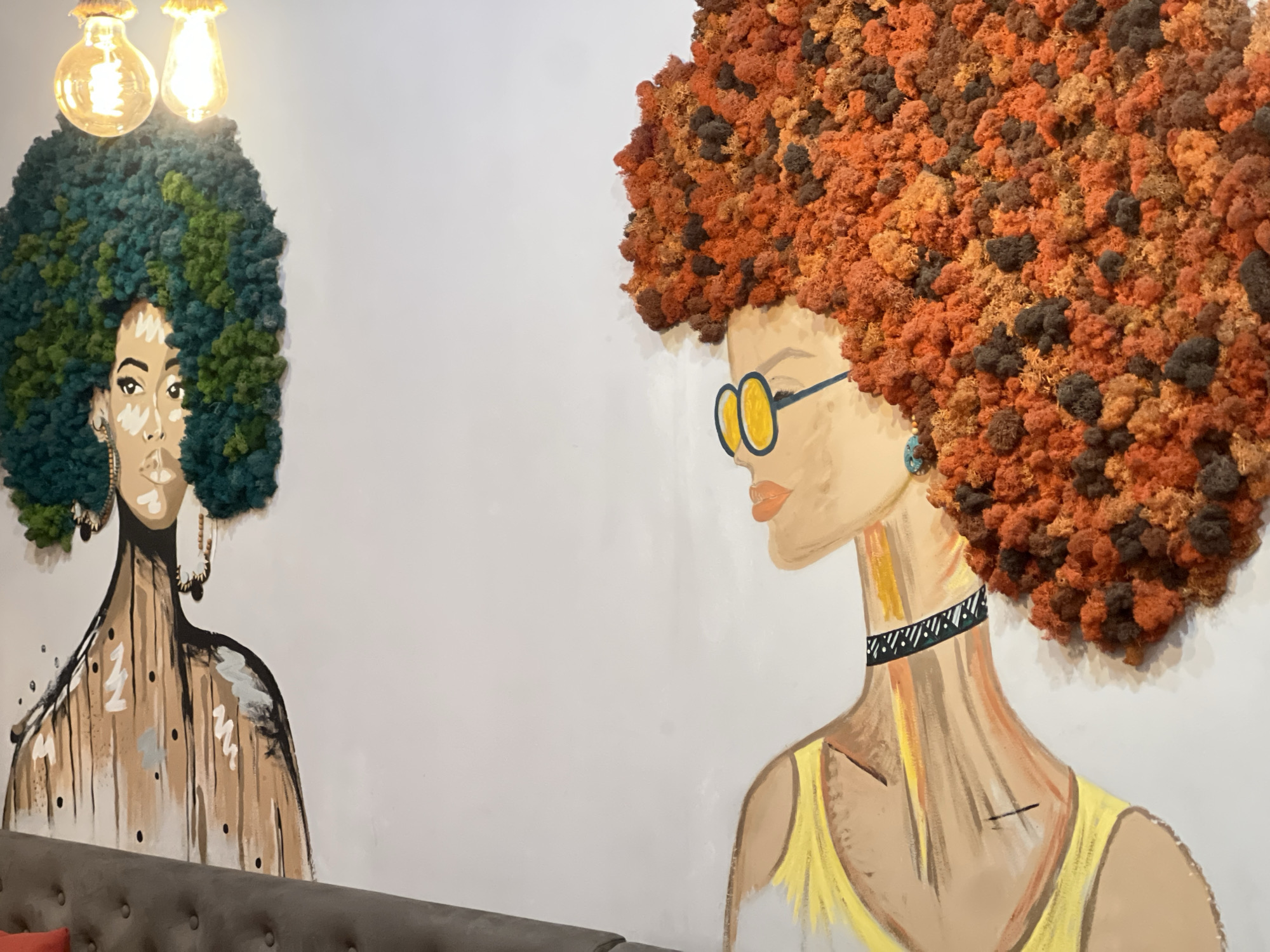 The image depicts an interior wall decorated with two mural-style paintings of women. The woman on the left has green textured hair reminiscent of moss or dense foliage, and a partially visible face with applied makeup. She wears large earrings. Her body is painted with broad, abstract brushstrokes in brown and white hues, suggesting a flowing garment. Above her hair are two illuminated light bulbs. The woman on the right has textured orange and brown hair. She wears yellow glasses and a patterned black and white choker. Her attire is painted in shades of yellow and white. The wall is painted a pale color, and a grey sofa with visible tufting runs along the bottom edge of the image. The exact location is unknown, but the prompt specifies the city of Yerevan, Armenia.