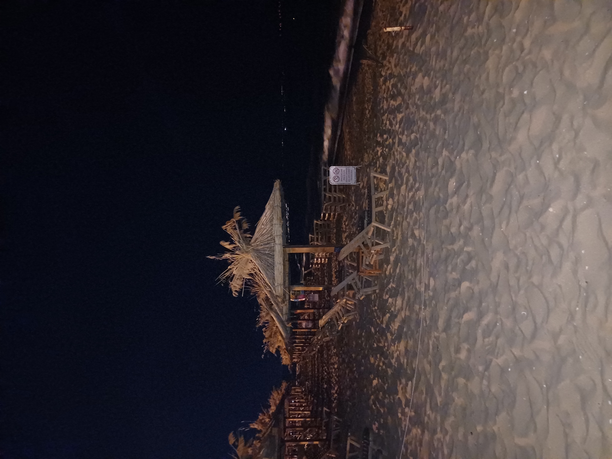The image depicts a beach at night. In the foreground, there is a sandy beach with numerous lounge chairs and thatched umbrellas lined up. The chairs are made of wood and appear to be empty. The umbrellas provide shade and are constructed from natural materials, possibly straw or palm fronds. The sand shows signs of being undisturbed, with footprints and the patterns of the waves.
In the middle ground, the sea is visible with gentle waves lapping at the shore. There are some distant lights on the horizon, suggesting the presence of a city or town. The sky is dark, indicating nighttime.
There is a sign with text and icons posted near one of the umbrellas. The text is not clearly legible due to the image quality and distance.
The scene suggests a quiet, possibly closed for the night, beach resort or public beach. Based on typical resort arrangements and the presence of umbrellas and lounge chairs, this could be a section of a beach along the Black Sea coast.
The city of Varna, Bulgaria, is a major coastal city on the Black Sea, known for its beaches and resorts. This image is consistent with the type of beach infrastructure found in Varna. Without more specific landmarks or identifying information within the image, it is not possible to pinpoint the exact location within Varna. However, the scene is characteristic of the Varna coastline.