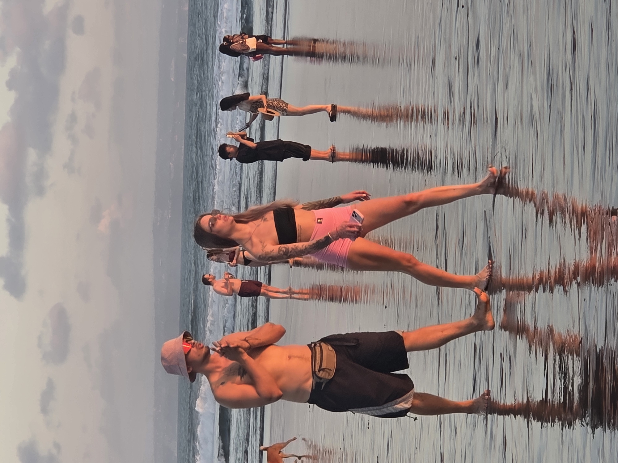 A group of people are walking on a wet sandy beach in North Kuta, Indonesia, during what appears to be sunset. In the foreground, a man wearing a bucket hat and sunglasses, and a woman in a strapless top and pink shorts, walk side-by-side. The man has a fanny pack around his waist. Behind them, several other individuals are visible, some wading in the shallow water and others standing on the shore. The ocean with waves is in the background, and the sky is filled with soft clouds. The wet sand reflects the light, creating a shimmering effect.