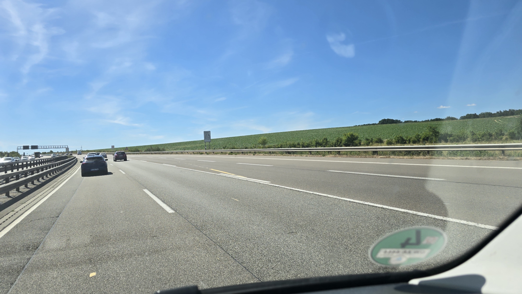 The image shows a highway in Kaiserslautern, Germany. The perspective is from inside a vehicle, looking forward. The highway has multiple lanes and is bordered by a guardrail on the left. Traffic is visible, with several cars driving ahead. On the right side of the highway, there is a large green field, possibly a crop field, extending up a gentle slope. The sky is blue with some scattered clouds. A road sign with directional arrows is visible on the left side of the highway. A green circular sticker with some text and symbols is present on the lower right portion of the windshield. The weather appears to be sunny and clear.