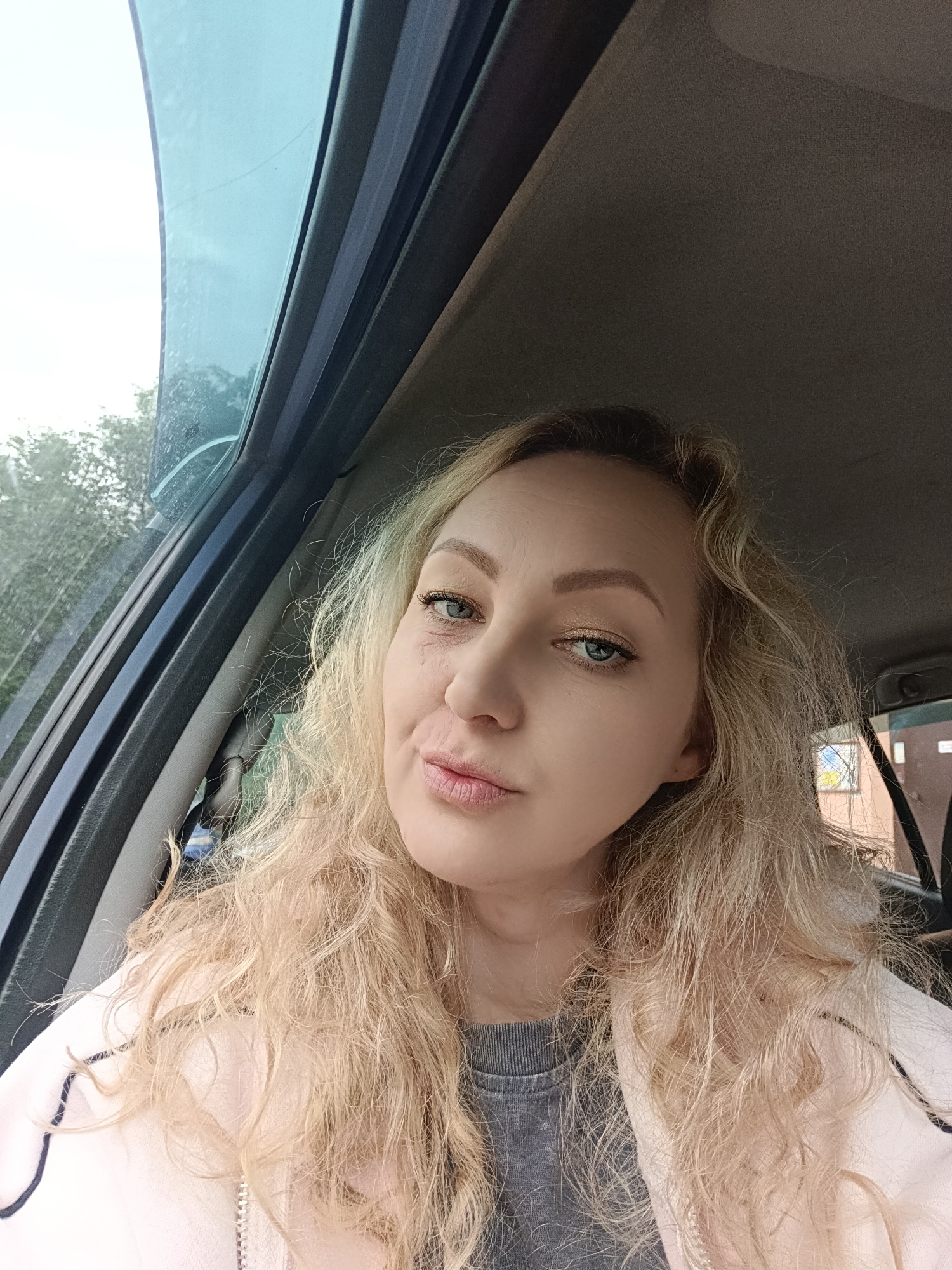 A medium close-up shot of a woman with light blonde curly hair sitting in a car. She has light skin, blue eyes, and is wearing eye makeup. A small scar or discoloration is visible under her left eye. She is wearing a grey shirt under a light pink jacket. The car's window is to her left, showing a glimpse of trees and an overcast sky. The interior of the car is visible, with a dark grey dashboard and window frame. The background is blurred, but a building can be partially seen through the rear window of the car. The location is implied to be Cherepovets, Russia.