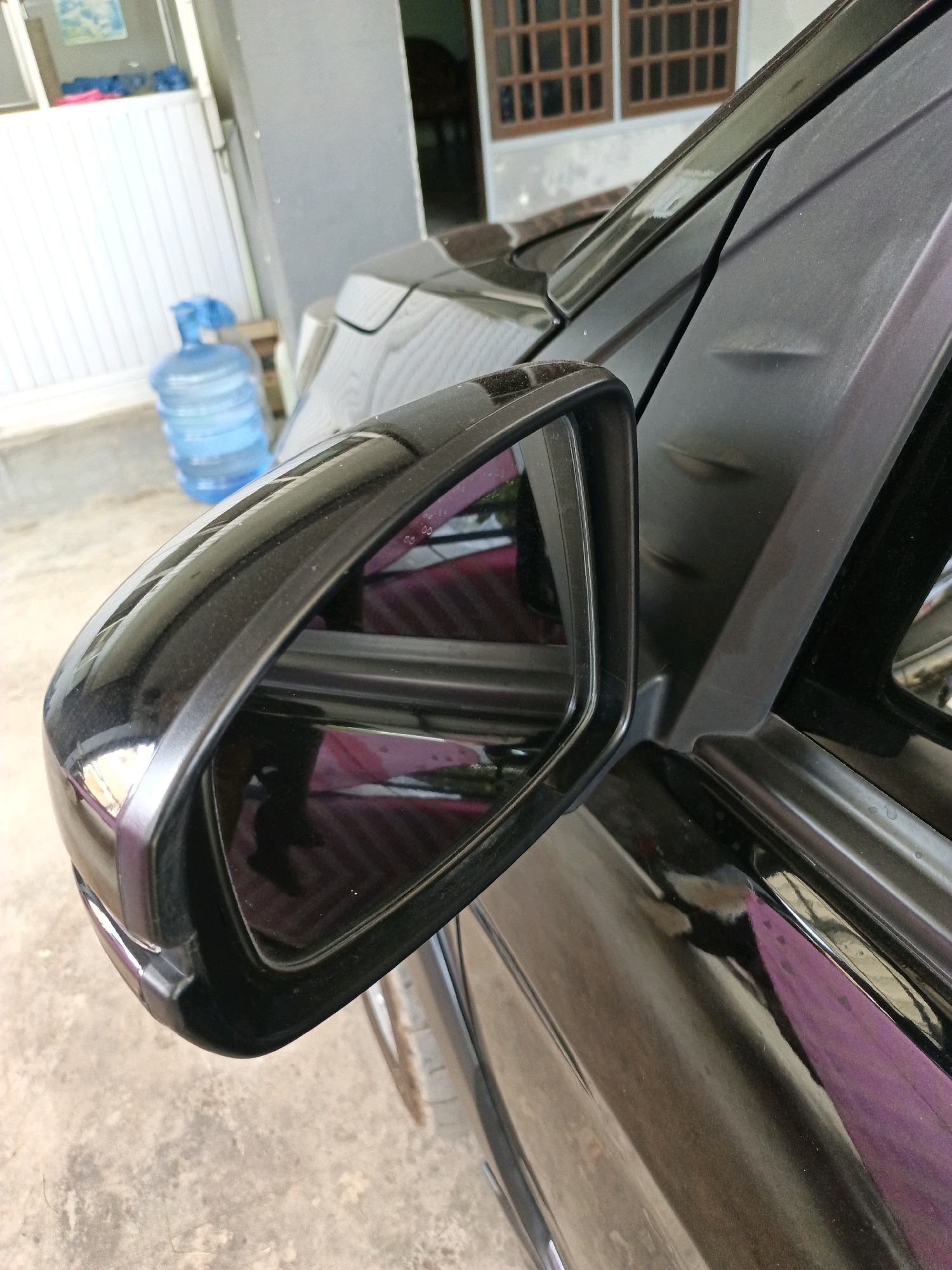 The image shows a close-up of a black car door and side mirror, likely parked outdoors on a concrete surface. In the background, a blue water cooler jug sits near a white slatted door. The car has a slightly dusty surface, and there appears to be a slight dent or imperfection on the black plastic trim of the car door. The side mirror reflects a purple and pink striped object. The scene appears to be in a residential or commercial area, possibly in Keleyan, Indonesia, based on the context provided. There are no people visible in the image.