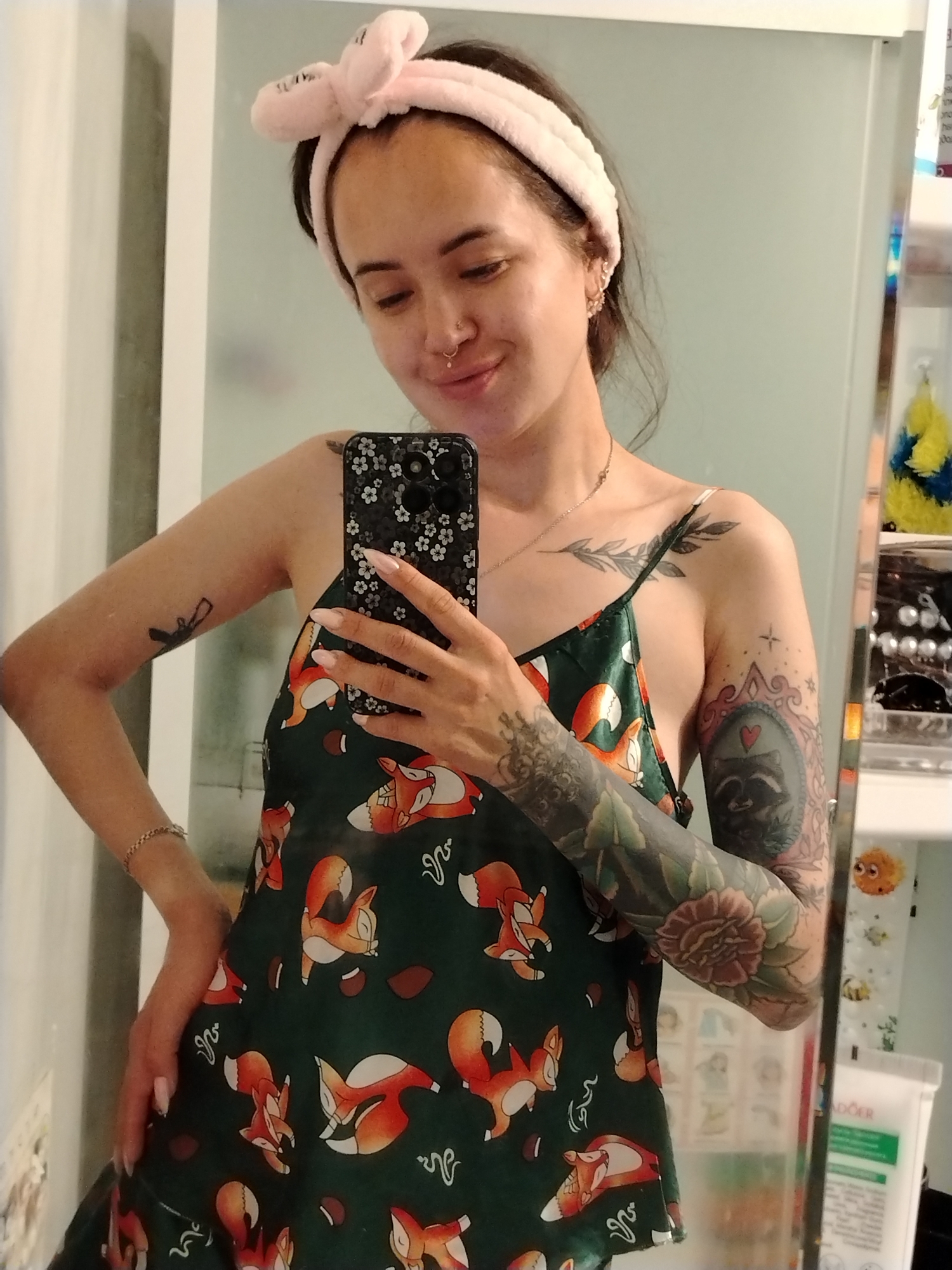 A young woman with dark hair and a light complexion is taking a mirror selfie in what appears to be a bathroom. She is wearing a green slip dress with a fox print and a pink fluffy headband. She has several tattoos on her arms and chest. Her right hand is holding a phone with a floral pattern case, and her left hand is on her hip. The background shows a mirror, a wall with vertical lines, and shelves with various items, including toiletries and decorative objects. The scene is indoors and well-lit, suggesting a personal living space. No specific location within Noviy, Russia can be determined from the visual information provided.