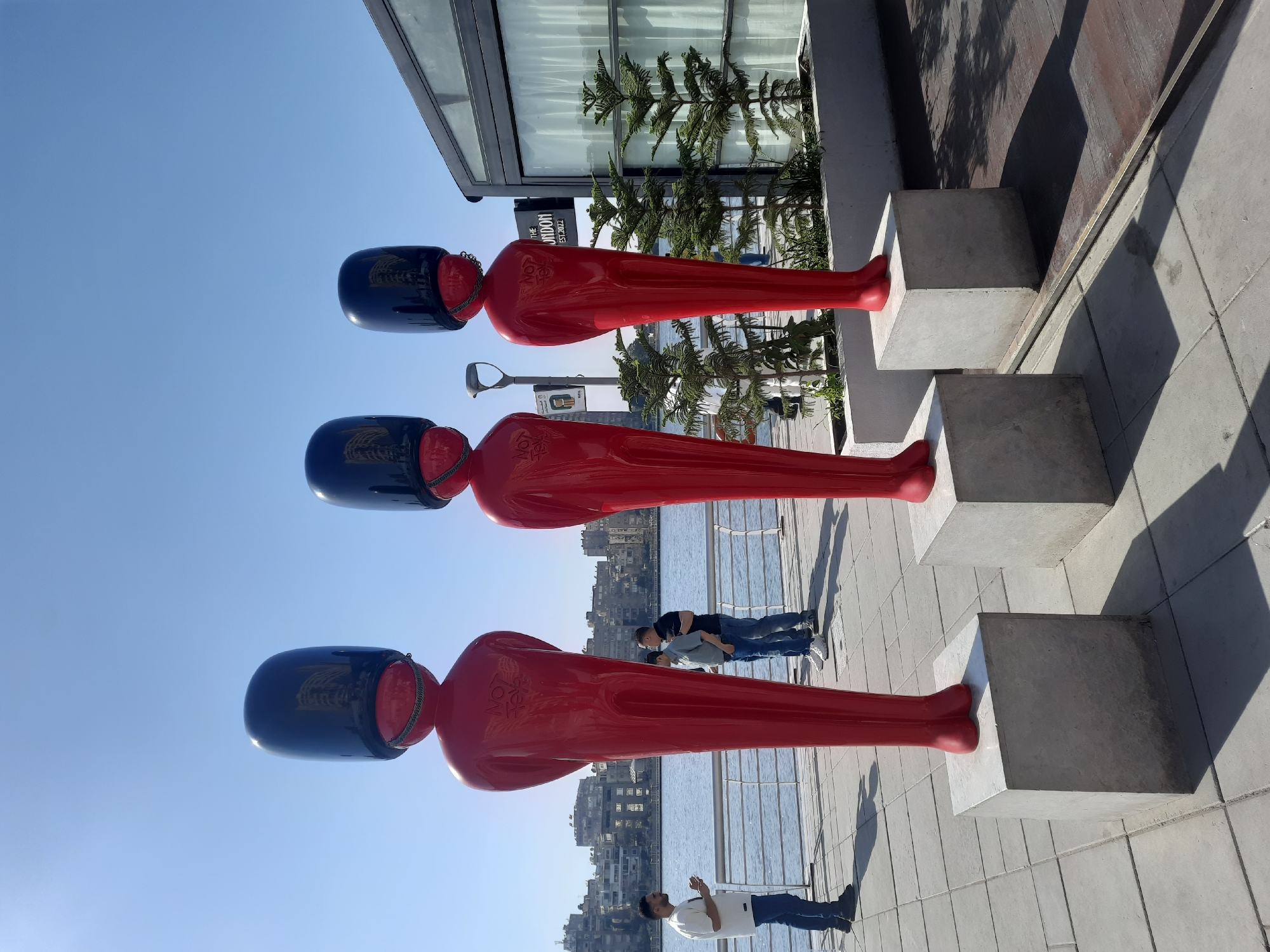 Three red, long, human-shaped sculptures, with black helmets, stand on concrete pedestals. People are nearby, on a concrete walkway beside a body of water reflecting a city skyline. A building with "THE LONDON" signage is behind the sculptures. The location is likely in Cairo, Egypt.