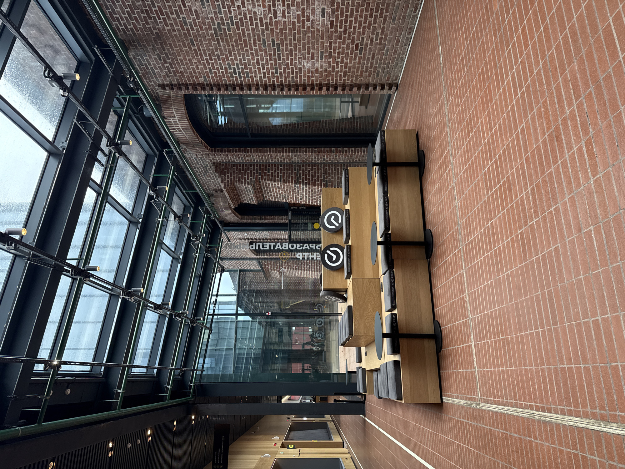 The image depicts an interior space with brick walls and flooring, featuring a modern design with large windows and metal framework. The lighting appears natural, coming from the exterior through the windows. There are wooden structures with seating, arranged along one side of the space. Signage with a circular symbol is visible on some of these structures. The scene is orderly and clean. The reflection in the windows shows external buildings, hinting at an urban location. Based on the visible Cyrillic text in the reflection ("Perm"), the city is Perm, Russia.