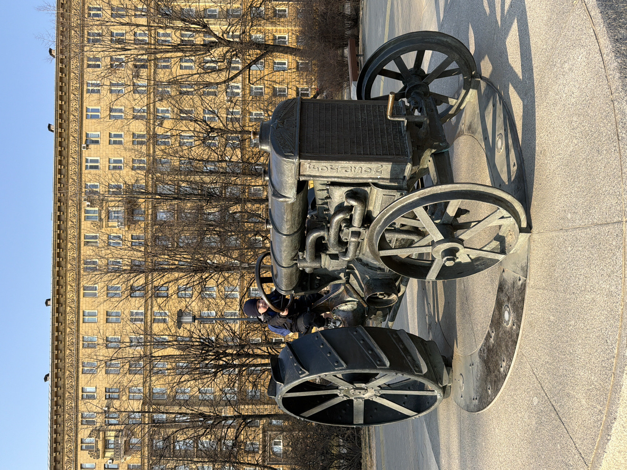 A low-angle, eye-level shot depicts an old, metal tractor statue situated on a circular concrete platform. A young child wearing a navy blue hat, jacket, and pants is seated on the tractor, holding the steering wheel.
The backdrop features a tall, light tan building with a multitude of windows arranged in a grid pattern. Bare tree branches partially obscure the building. The sky is light blue.
The tractor is the primary focus of the image. The surrounding environment suggests the location is Saint Petersburg, Russia. The circular concrete platform the tractor sits on is gray. The shadow of the tractor is cast onto the concrete platform.