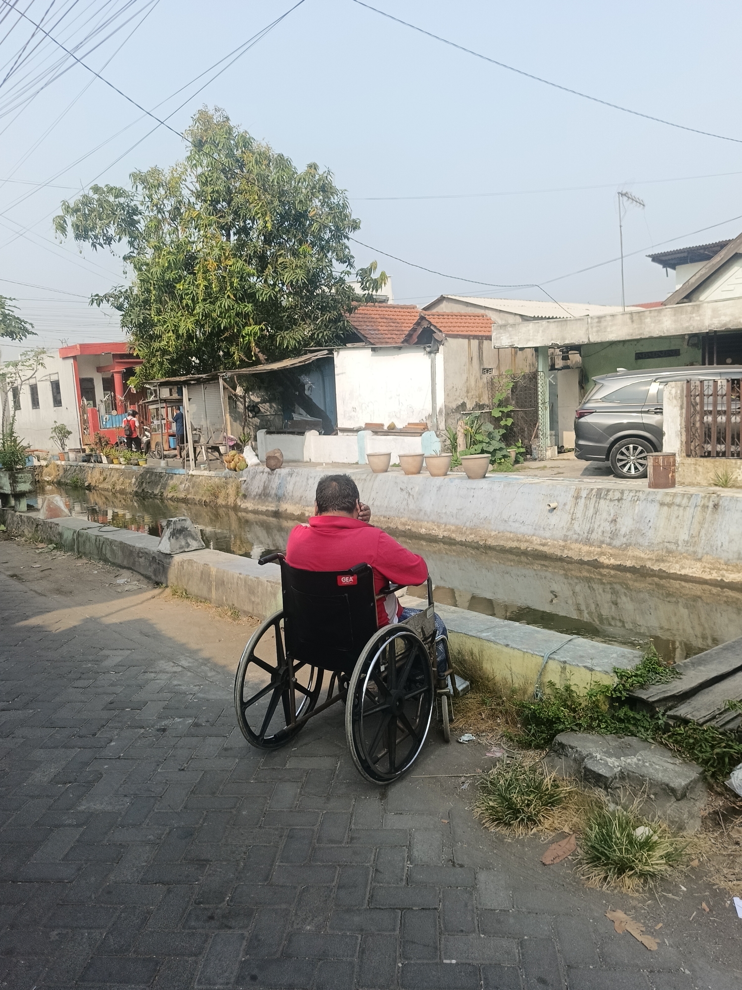 A man in a red shirt is sitting in a wheelchair facing away from the camera, by a canal. The wheelchair is black and has large wheels. The man appears to be looking at the canal. In the background, there are houses, a tree, and a gray car. The scene is outdoors, under a clear sky, suggesting a sunny day. The location is identified as Gedangan, Indonesia. The canal has concrete walls, and the walkway beside it is paved with interlocking bricks. There are some plants growing along the canal bank and on the walkway. A small structure with a corrugated roof is also visible near the canal, and there are potted plants in front of the houses.