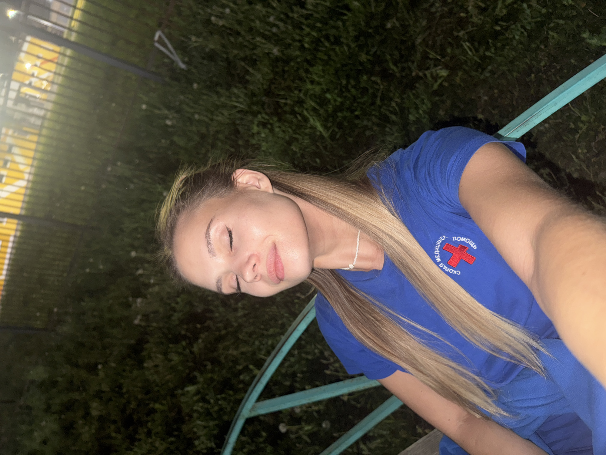 A young, blonde woman is the subject of this medium shot, taken outdoors at night. She is slightly turned away from the camera and looking down with her eyes closed. Her skin is fair. She is wearing a blue t-shirt, upon which is embroidered a red cross and Cyrillic text stating "СКОРАЯ МЕДИЦИНСКАЯ ПОМОЩЬ," which translates to "Emergency Medical Assistance" in English. A small, white necklace is visible on her neck.
The woman is sitting on a park bench with blue-green, metallic support bars. The backdrop appears to be foliage and grass, suggesting a park or green space. In the distant left, there are some faint lights and hints of urban infrastructure, indicating the setting could be in or near a city.
While it is claimed this location is Chaikovsky, Russia, that information is not visible in the provided image. The visual information alone does not confirm this location.