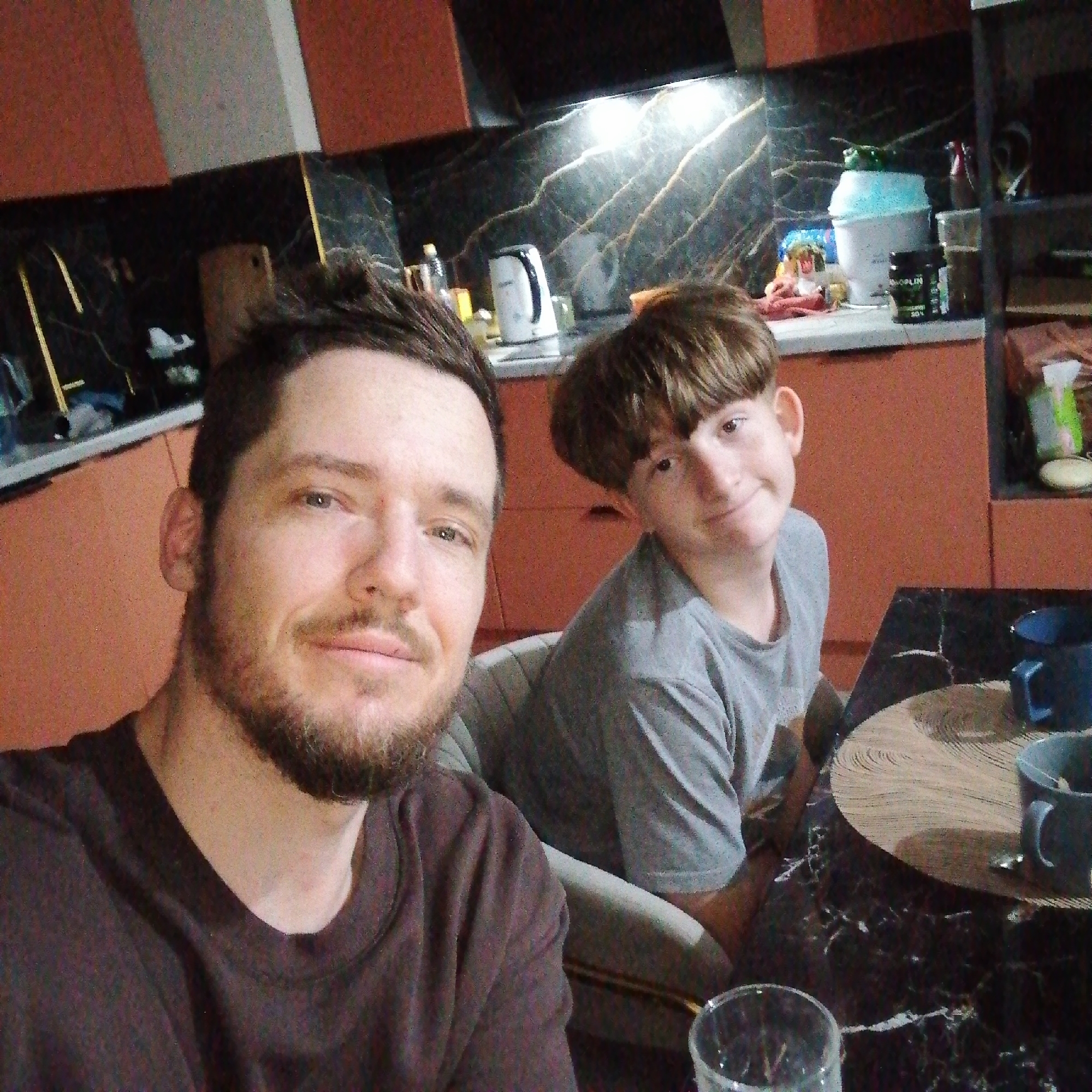 The image captures two individuals, a man and a boy, seemingly in a domestic kitchen setting. The man, in the foreground, appears to be taking a selfie, as he faces the camera with a slight smile. He has short, dark hair and a beard. The boy is seated slightly behind and to the right of the man, also facing the camera with a subtle smile. He has a distinctive bowl-shaped haircut.
The background reveals a modern kitchen with orange cabinets and a black, marbled backsplash. Visible kitchen appliances include a kettle and various containers. A circular wooden placemat and a blue mug sit on a black marble table. A glass tumbler is also on the table in the foreground.
The location and city are unknown. The environment is domestic and well-lit.