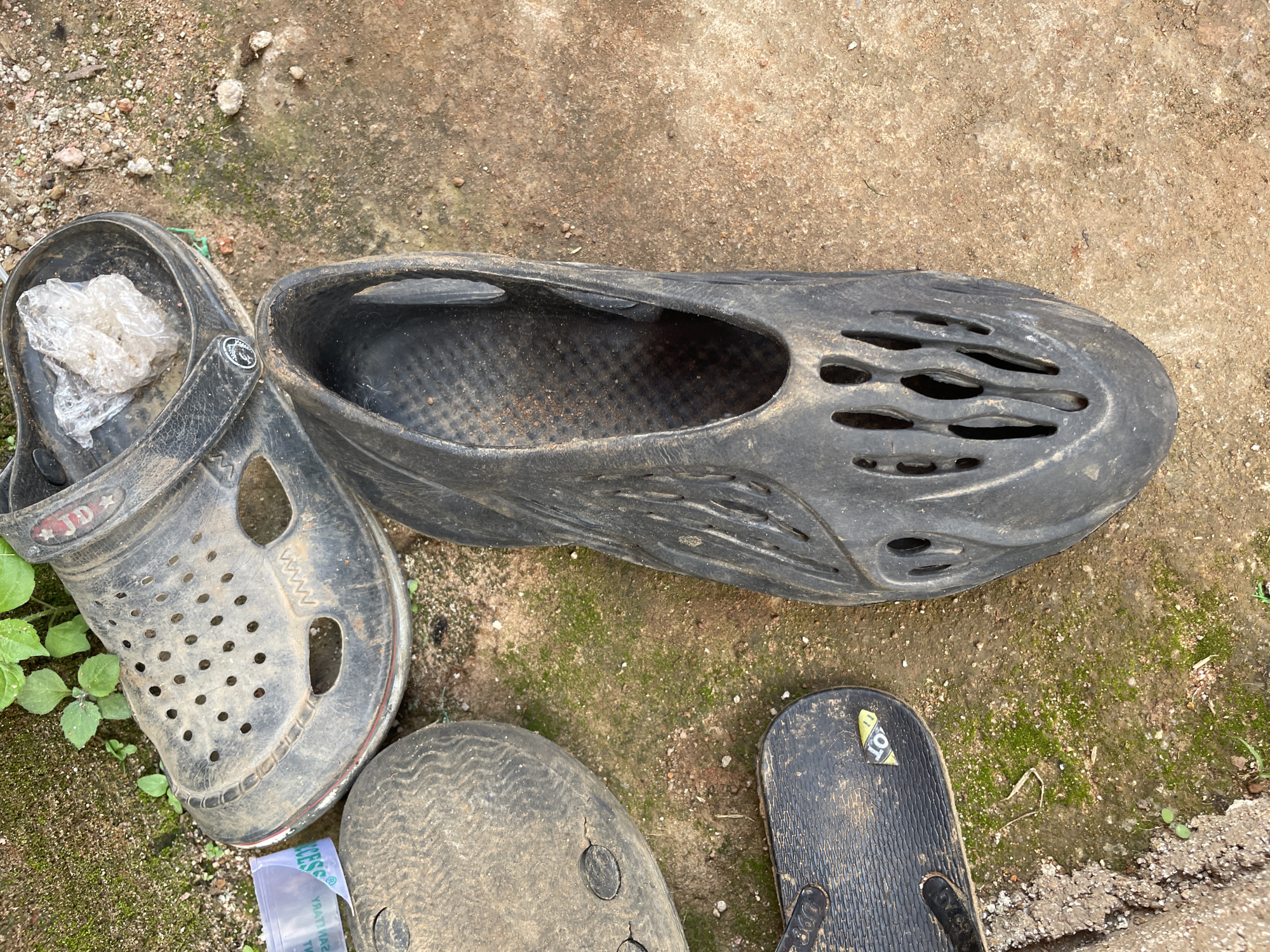 The image displays a collection of dirty footwear on a mix of soil and concrete. In the upper left quadrant, a black clog-style shoe, appearing somewhat worn, is visible, with a crumpled piece of clear plastic inside. To the left of this clog is a portion of another sandal, its sole textured and dusty. In the lower left quadrant, a black flip-flop with a textured sole is laid out. The largest object in the image, situated in the right half, is a black, molded shoe with an open, ventilated design. The ground appears to be a mixture of dirt, small stones, and patches of moss, suggesting an outdoor environment. The scene is likely located in Durimi, Nigeria, as per the prompt, indicating a possibly dusty and humid climate that contributes to the state of the footwear and ground cover. No people are visible in the image.