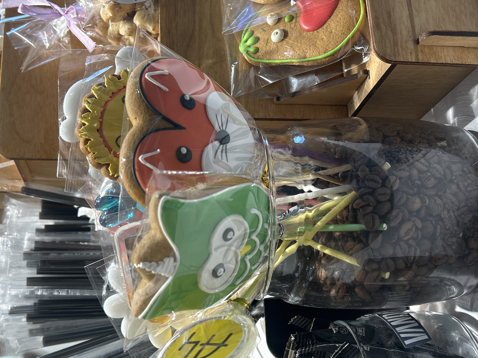 This close-up image, likely taken in a shop or market in Tolyatti, Russia, showcases a vibrant display of packaged artisanal food items. Dominating the center-right are several elaborately decorated gingerbread cookies, each individually sealed in clear plastic. A prominent cookie is shaped like a red animal head, possibly a fox or squirrel, featuring white snouts, black eyes, and delicate white icing details. Below it, a green owl-shaped cookie is visible, characterized by large white eyes and decorative green and white icing. To the left of the red animal cookie, a bright yellow sunflower-shaped cookie is displayed. Further left, another clear bag contains smaller, light-colored cookies or treats, tied with a delicate purple ribbon.
In the background, on the right side of the frame, a large clear glass container is filled with dark roasted coffee beans, from which several thin yellow and green decorative sticks or stirrers emerge. To the far left, stacks of individually wrapped black plastic straws or stirrers are neatly arranged. The products are presented on light brown wooden display shelving, suggesting a crafted or rustic aesthetic. The scene is brightly lit, indicating an indoor setting during the daytime. Visible text includes partial Cyrillic characters "ЧЧ" on a yellow sticker attached to the owl cookie's packaging, and parts of an "EAC" conformity mark and striped patterns on a black package at the bottom.