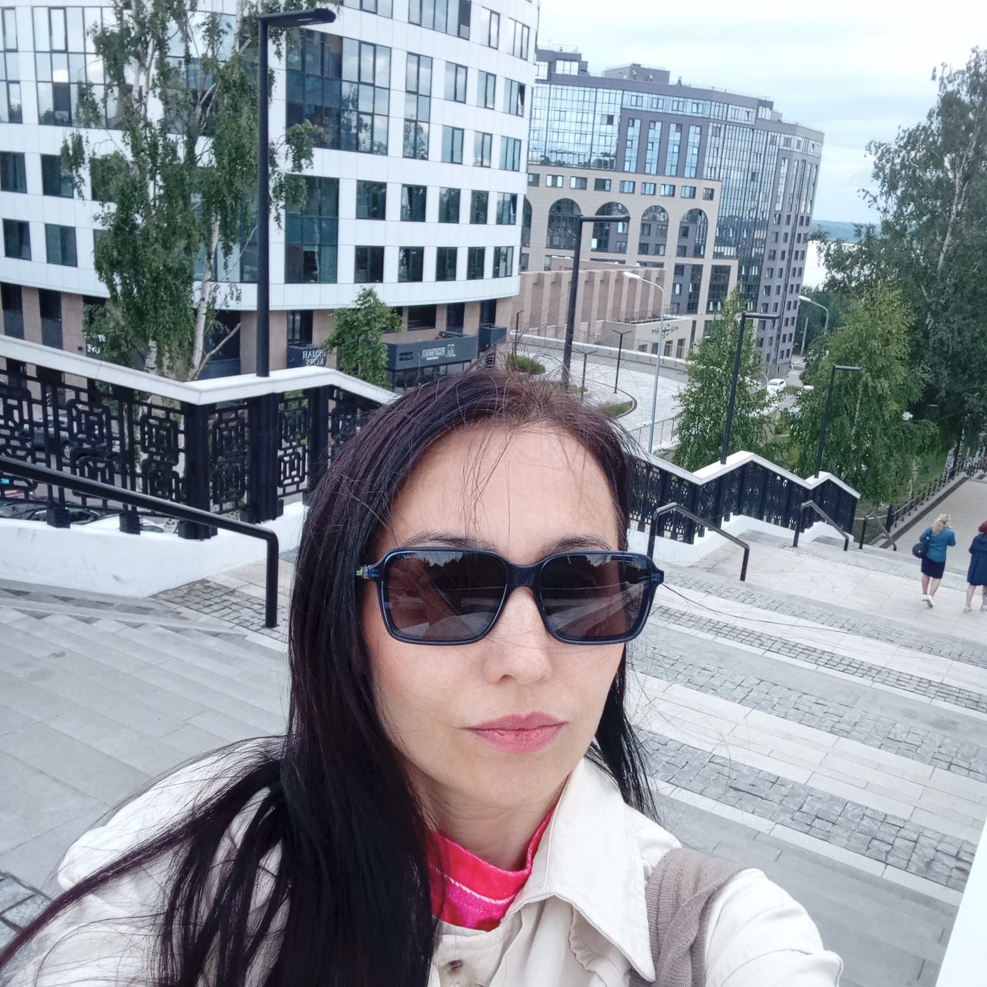 A woman with long, dark hair and dark sunglasses is captured in a selfie perspective, positioned in the foreground and center of the image. She wears a light-colored collared shirt over a pink garment and a light brown vest. Her sunglasses bear the visible brand name "DIOR" on the right arm. She is standing on a wide, multi-tiered staircase constructed with light and dark grey paved stones, which extends into the midground and background. The staircase features black metal railings with geometric patterns, topped with white handrails.
In the midground and background, the scene reveals a modern cityscape in Izhevsk, Russia. To the left, a multi-story building with a curved white facade and numerous rectangular windows is prominent. Adjacent to it on the right stands another multi-story grey building featuring both large glass sections and distinct arched windows on its lower levels. Below these buildings, signs in Cyrillic script are visible for businesses such as "НАИР" and "ДИМОНЕТ СС". Several deciduous trees, including a tall birch, are interspersed among the buildings and along pathways.
Further into the background, the staircase descends, leading to a visible road with a white car and more trees. Two other individuals, appearing to be women, are walking down the stairs in the distant midground on the right. Lampposts are strategically placed along the pathways and staircase. The distant horizon shows a hazy outline suggesting a large body of water, likely the Izhevsk Pond, under an overcast sky. The overall environment indicates a developed urban area with modern architecture and public recreational spaces.