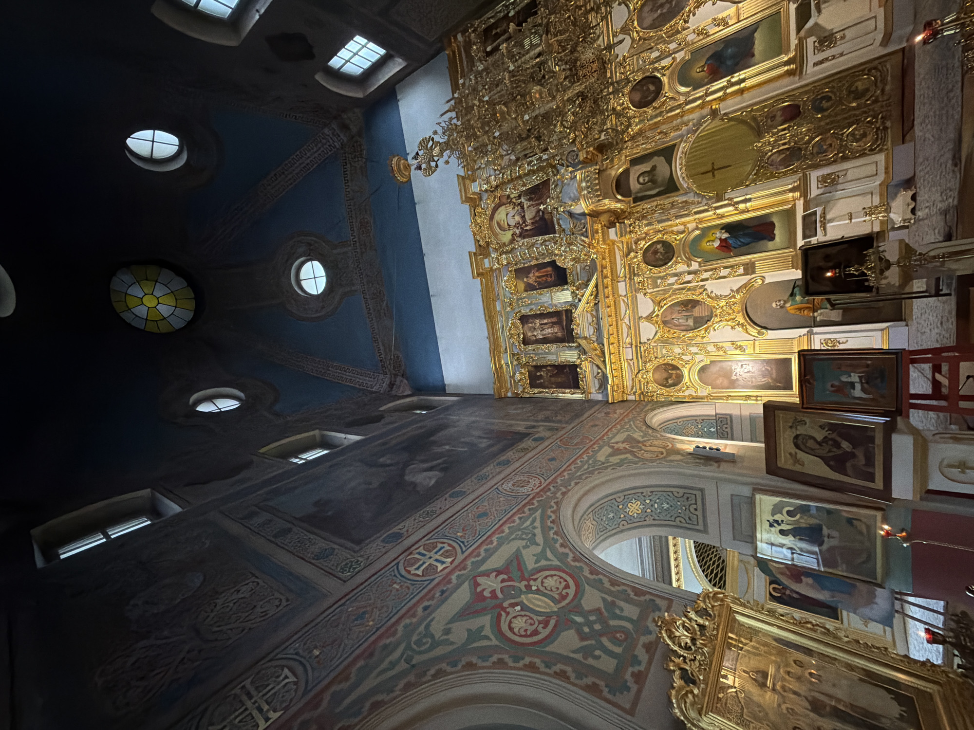 The image shows the interior of a church, likely the Kazan Cathedral in Saint Petersburg, Russia, given the ornate gold iconostasis and the architectural style. The camera angle is low and wide, looking up towards the ceiling and the altar area.
In the foreground on the left, a large, ornate gold-framed icon is visible. To its right, several other icons are arranged on stands, some propped against each other. One icon depicts a religious scene with figures, another shows a seated figure, and a third features a woman in a dark robe. A single red candle in a candlestick burns in front of one of these icons.
The central and right portions of the image are dominated by the magnificent gold iconostasis, a screen adorned with numerous religious paintings set within intricately carved golden frames. Many of these paintings depict religious figures, including Christ and Mary. The iconostasis features large double doors, known as the Royal Doors, at its center. Above the iconostasis, a large, ornate golden chandelier hangs from the ceiling.
The walls and ceiling of the church are decorated with frescoes and intricate patterns in shades of blue, red, gold, and beige. Several windows are visible, including a distinctive circular stained-glass window in the ceiling, as well as rectangular windows with multi-pane designs. The ceiling is a deep blue, with arched sections and decorative moldings.
The overall impression is one of grandeur and religious solemnity, with the rich gold accents and detailed artwork filling the space. The lighting appears to be a mix of natural light from the windows and artificial light from the chandelier. There are no people clearly visible in the image.