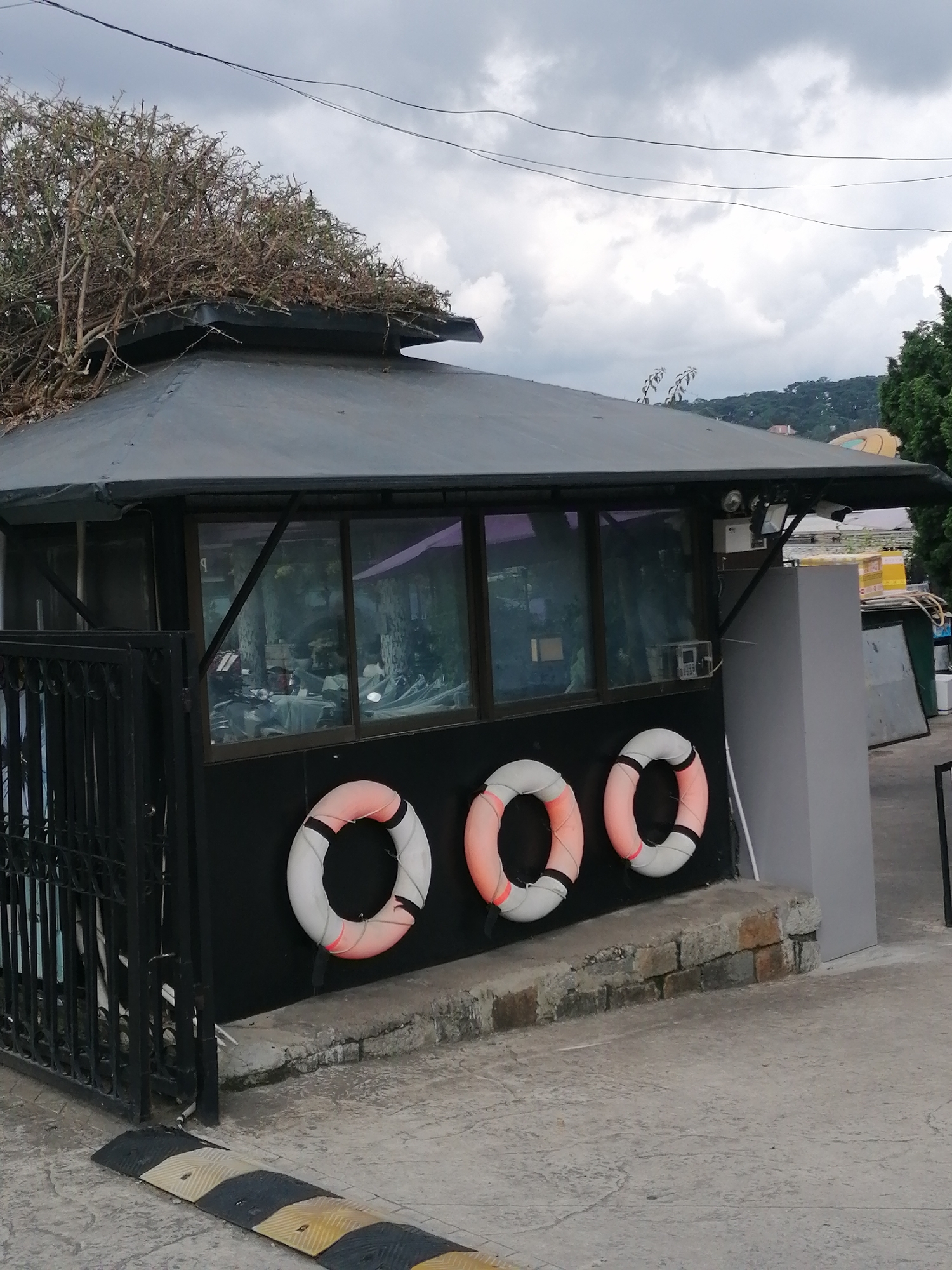 A dark grey, multi-sided structure with a slanted roof is visible, partially obscured by a dense clump of dry branches and twigs on its top. The structure features large windows on one side, reflecting the surroundings and revealing tables and chairs within. Three life preservers, white with orange accents, are mounted on the dark wall of the structure. A black metal gate is positioned to the left. In the foreground, a yellow and black speed bump is laid across a grey concrete surface. The background shows a grey, overcast sky, some greenery on a hillside, and hints of buildings. The overall scene appears to be an outdoor area, possibly near a body of water given the life preservers, and is located in Da Lat, Vietnam.
