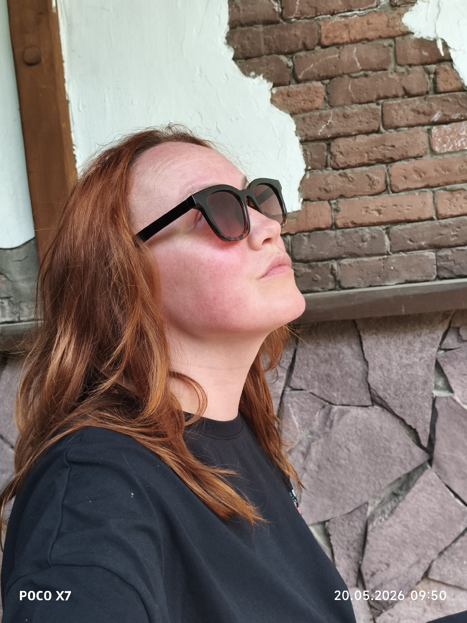 A fair-skinned woman with reddish-brown hair wears black-framed sunglasses and a black t-shirt. She is looking up and to her right. The background consists of a brick wall with patches of white plaster and a lower portion of stacked stone slabs. The image includes a timestamp and camera model label: "POCO X7 20.05.2026 09:50." The location is Chaikovsky, Russia.